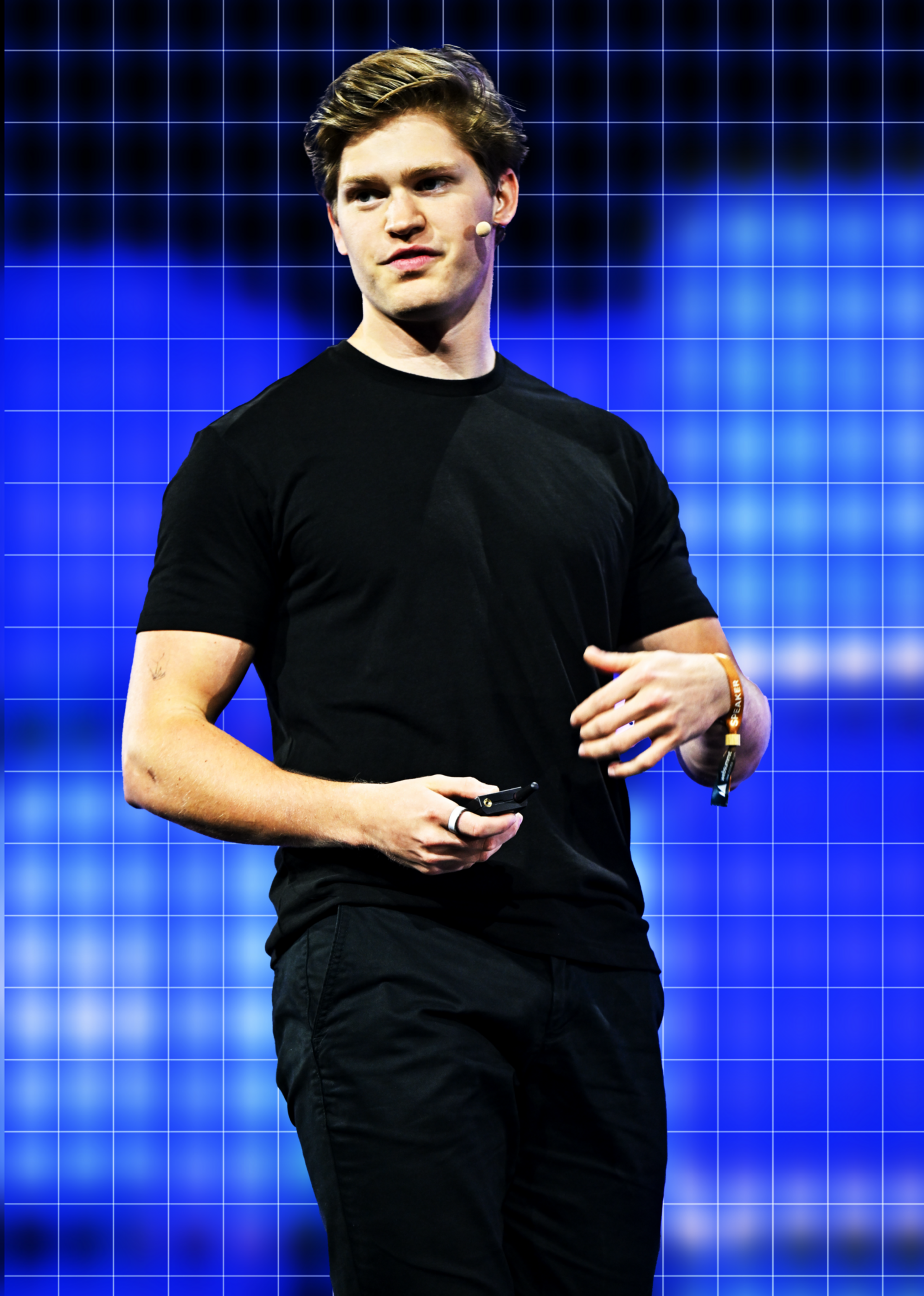 A young man with a headset mic wears a black t-shirt and pants, standing in front of a blue, grid-like digital background, holding a small device.