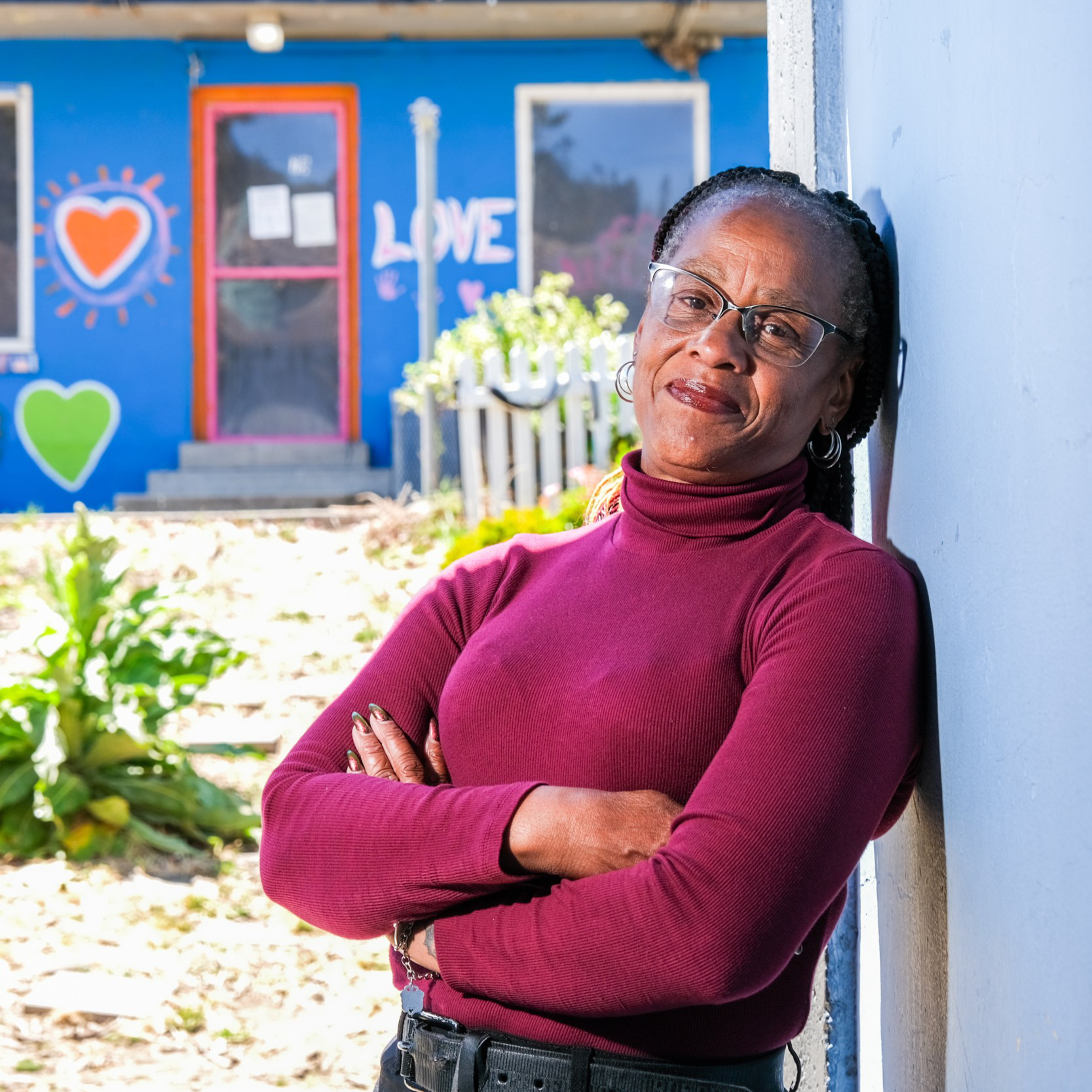 A woman in a burgundy turtleneck, wearing glasses, leans against a wall with her arms crossed. The background shows a blue building with colorful heart and love decorations.