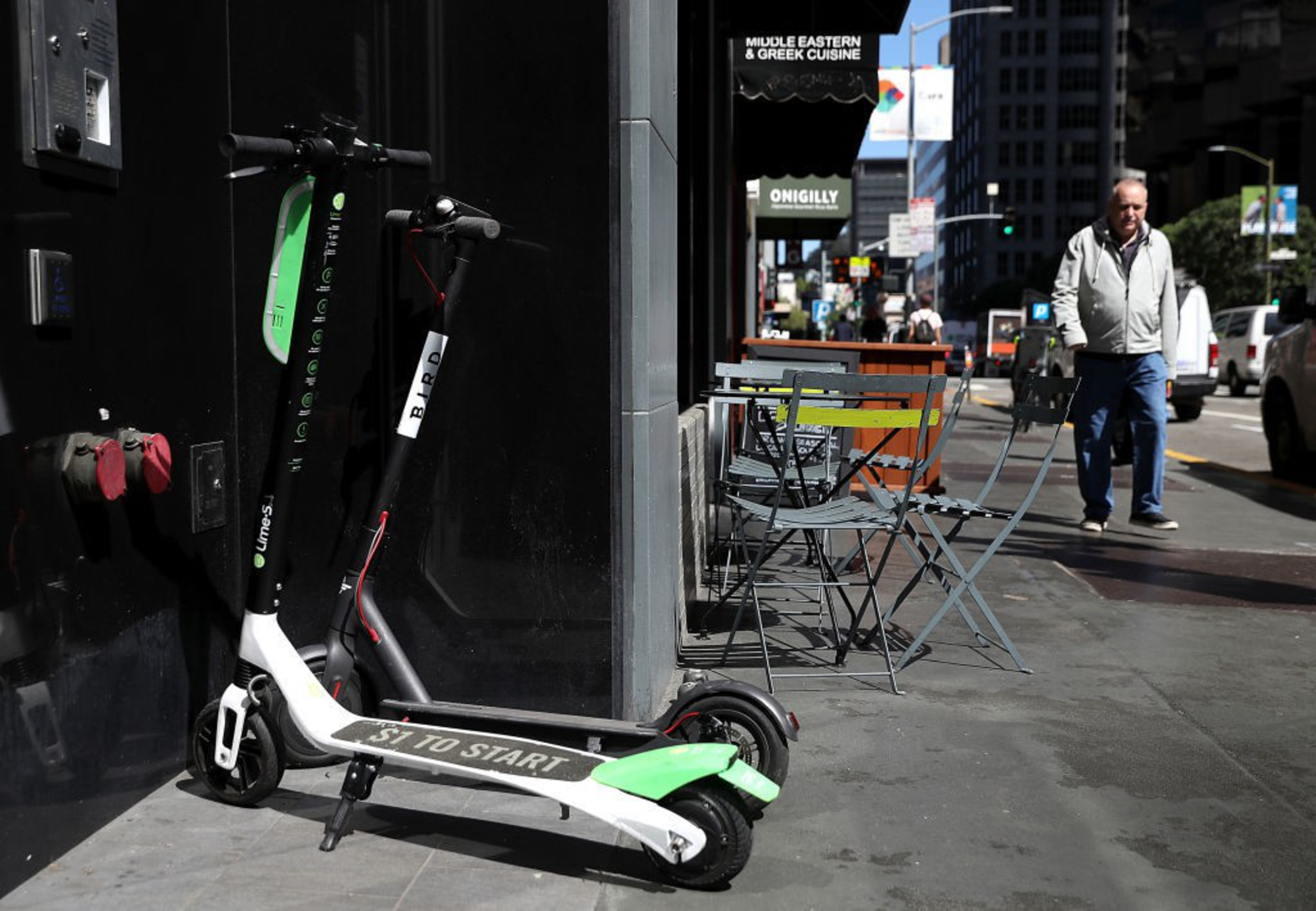 Two parked electric scooters lean against a black wall near a sidewalk café with empty tables and chairs, while a man walks down the street.