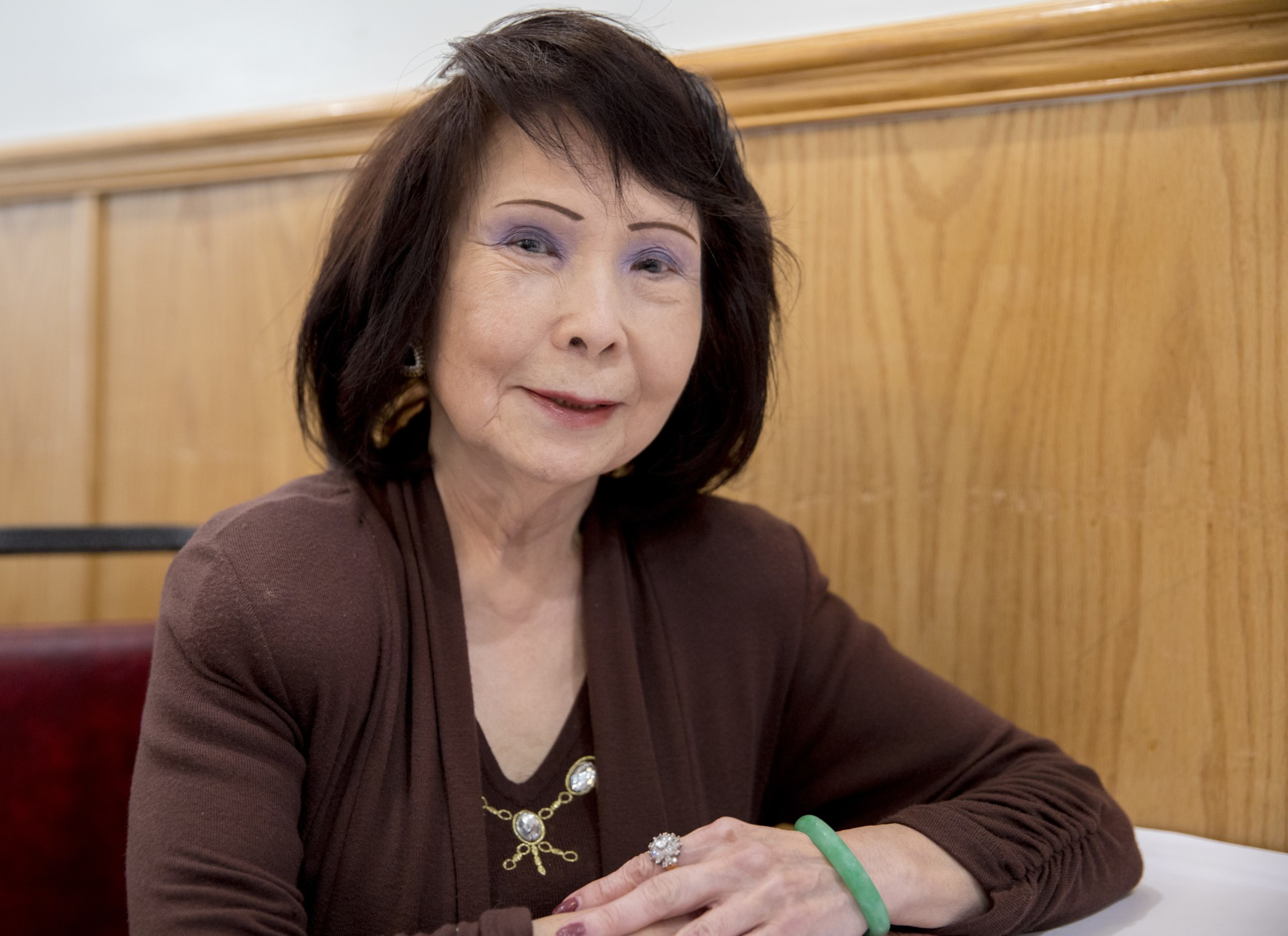 A woman with dark hair sits smiling, wearing a brown top with a decorative brooch. She has a green bracelet and a ring, and there's wood paneling behind her.