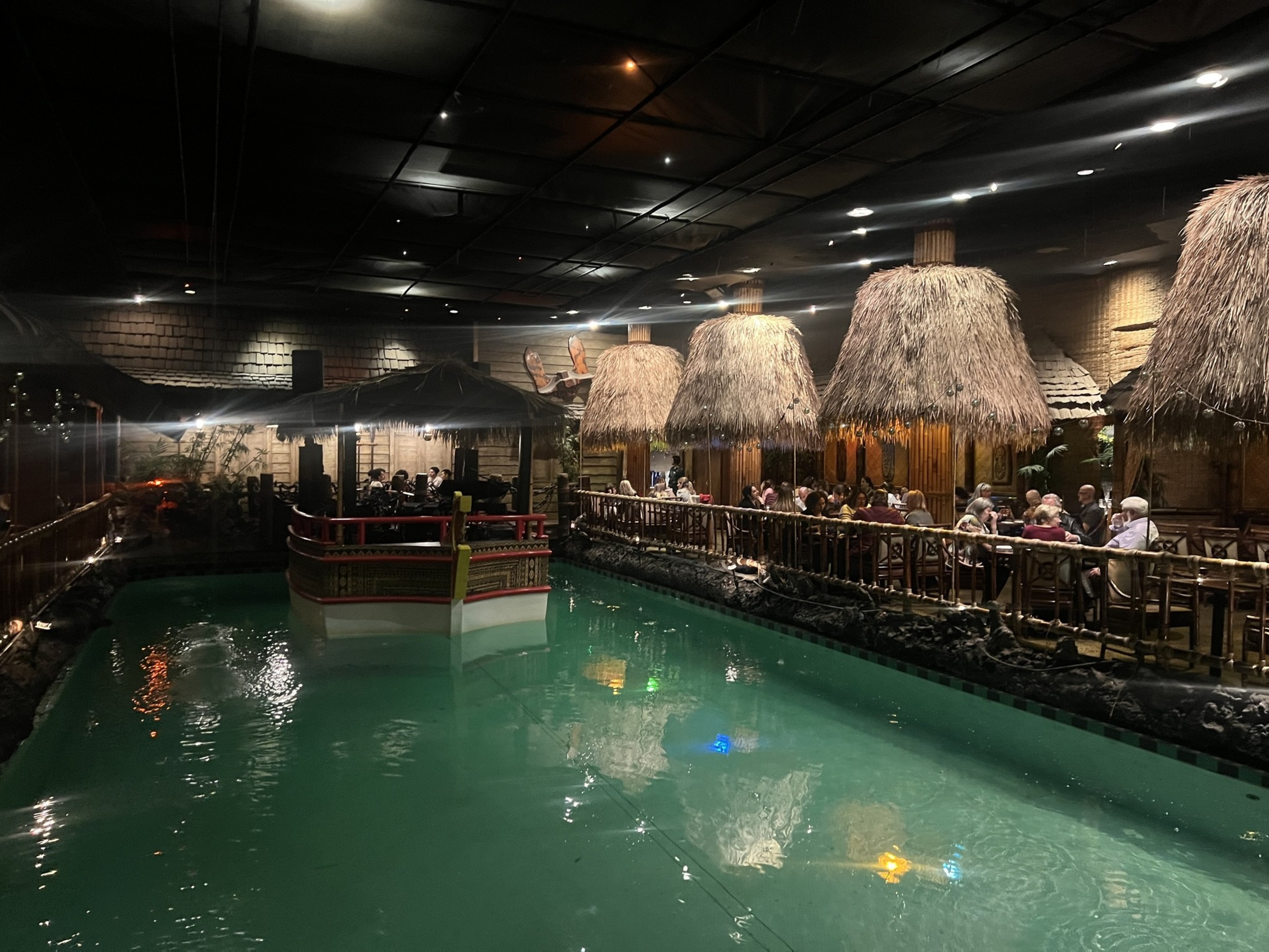 A swimming pool in a restaurant with thatched roof tables around it. 