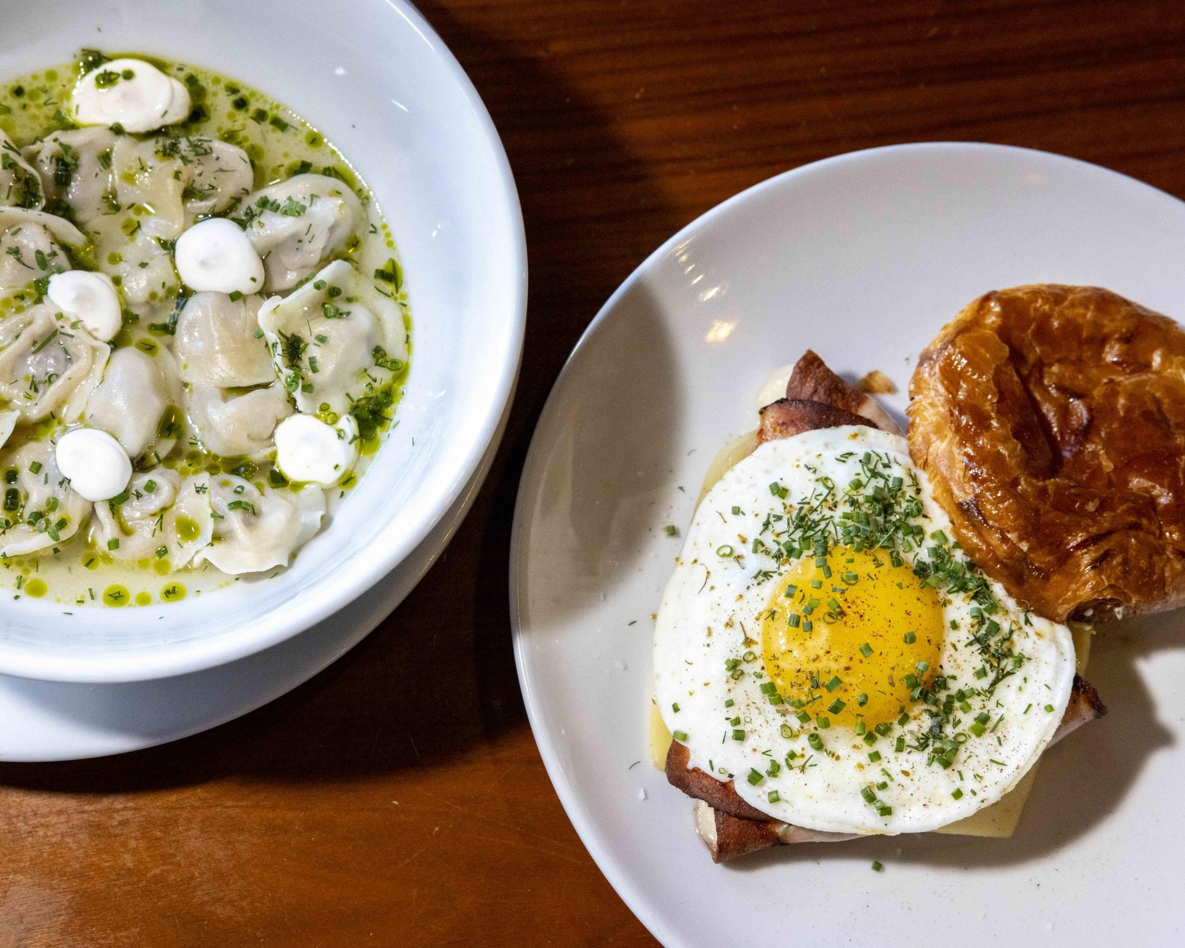 The image shows a plate of dumplings in broth with herbs and dollops of cream, next to a dish featuring a sandwich topped with a fried egg and a flaky pastry.