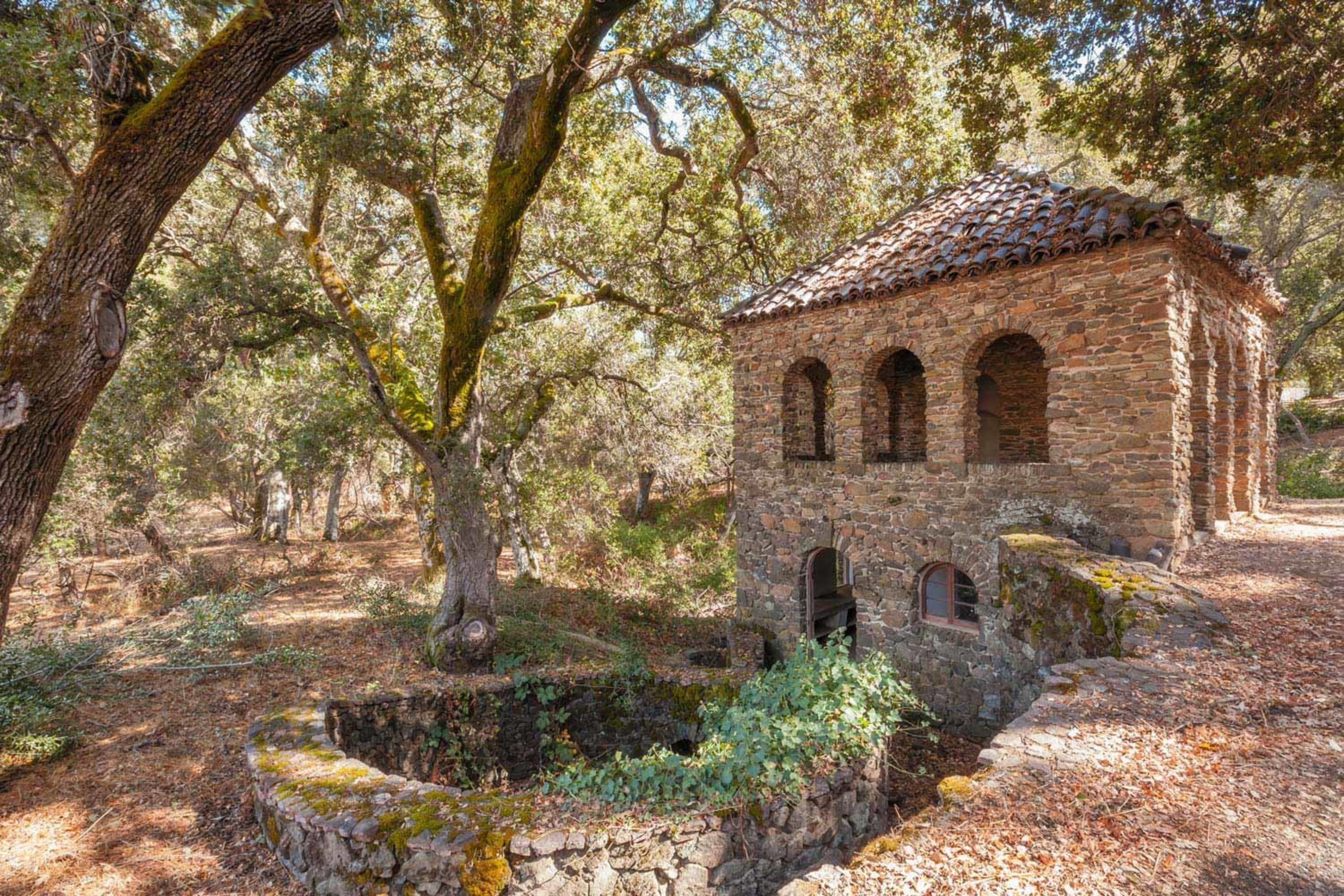 A small stone building with arched windows and a tiled roof sits beside a circular stone structure, surrounded by tall trees and dry leaves on the ground.