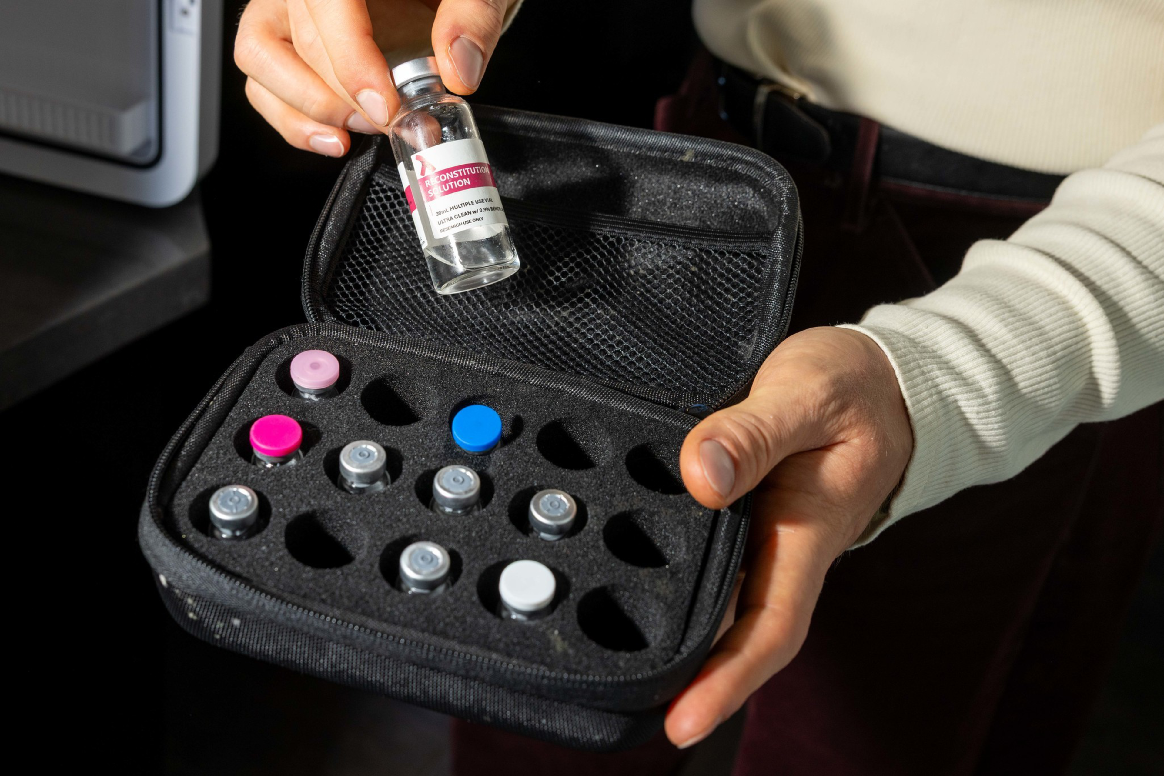 A person holds a black case with several small vials inside, one vial labeled “CONSTITUTIONAL SOLUTION” is held separately in the other hand.