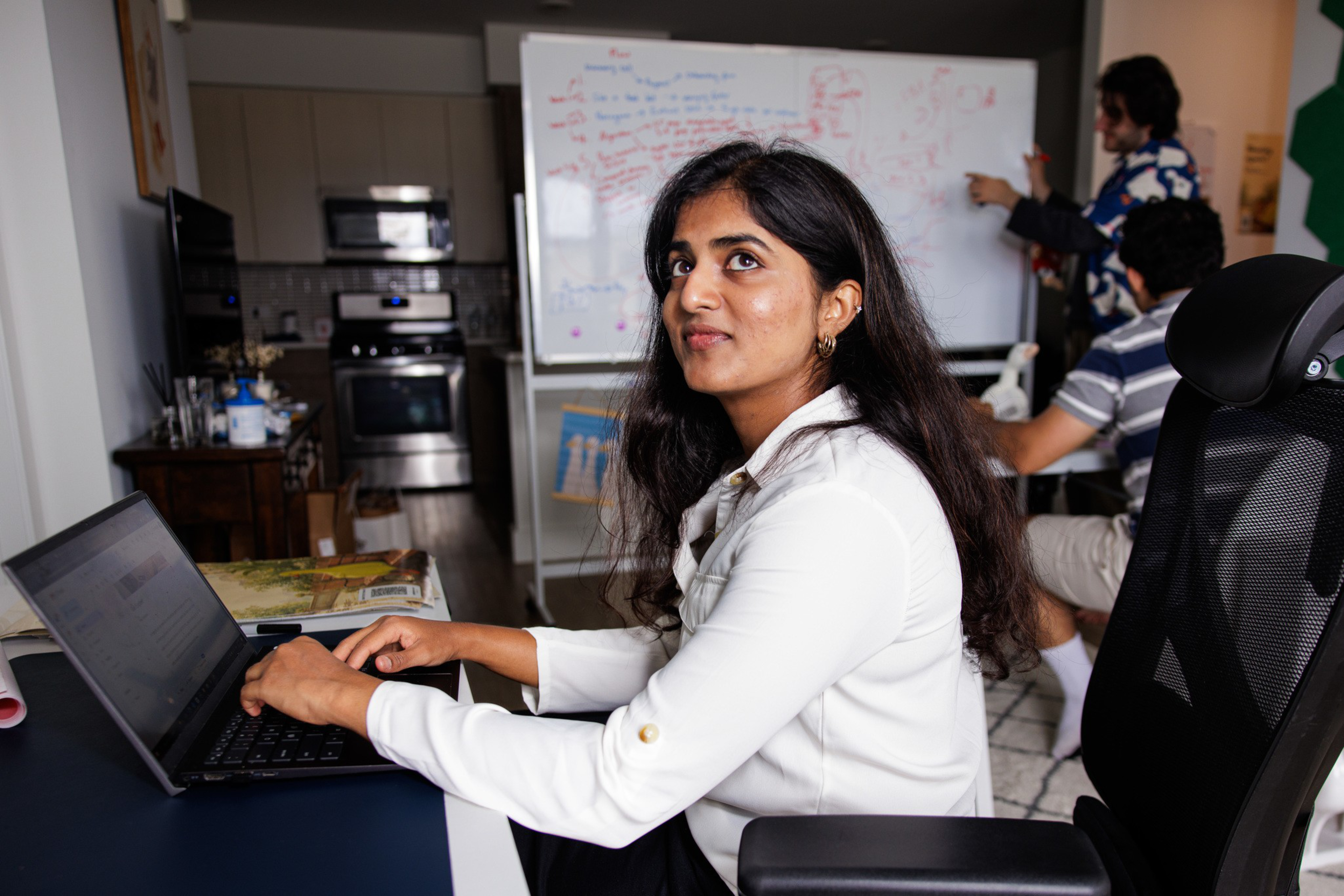 A woman in a white shirt sits at a laptop, while two men discuss notes on a whiteboard in a room with a kitchen in the background.