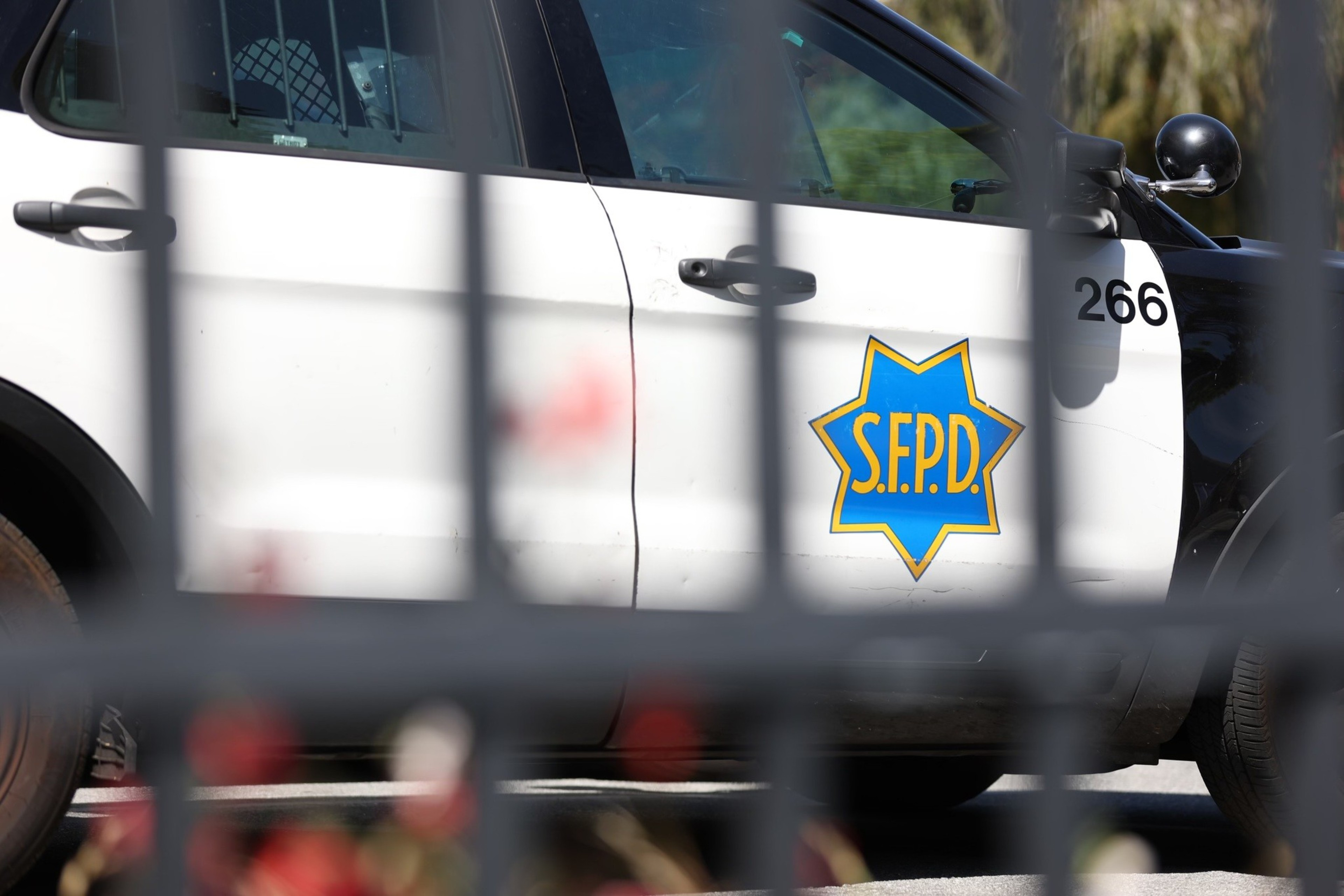 A white and black San Francisco Police Department car with the S.F.P.D. badge and the number 266 on the front door is seen behind a metal fence.