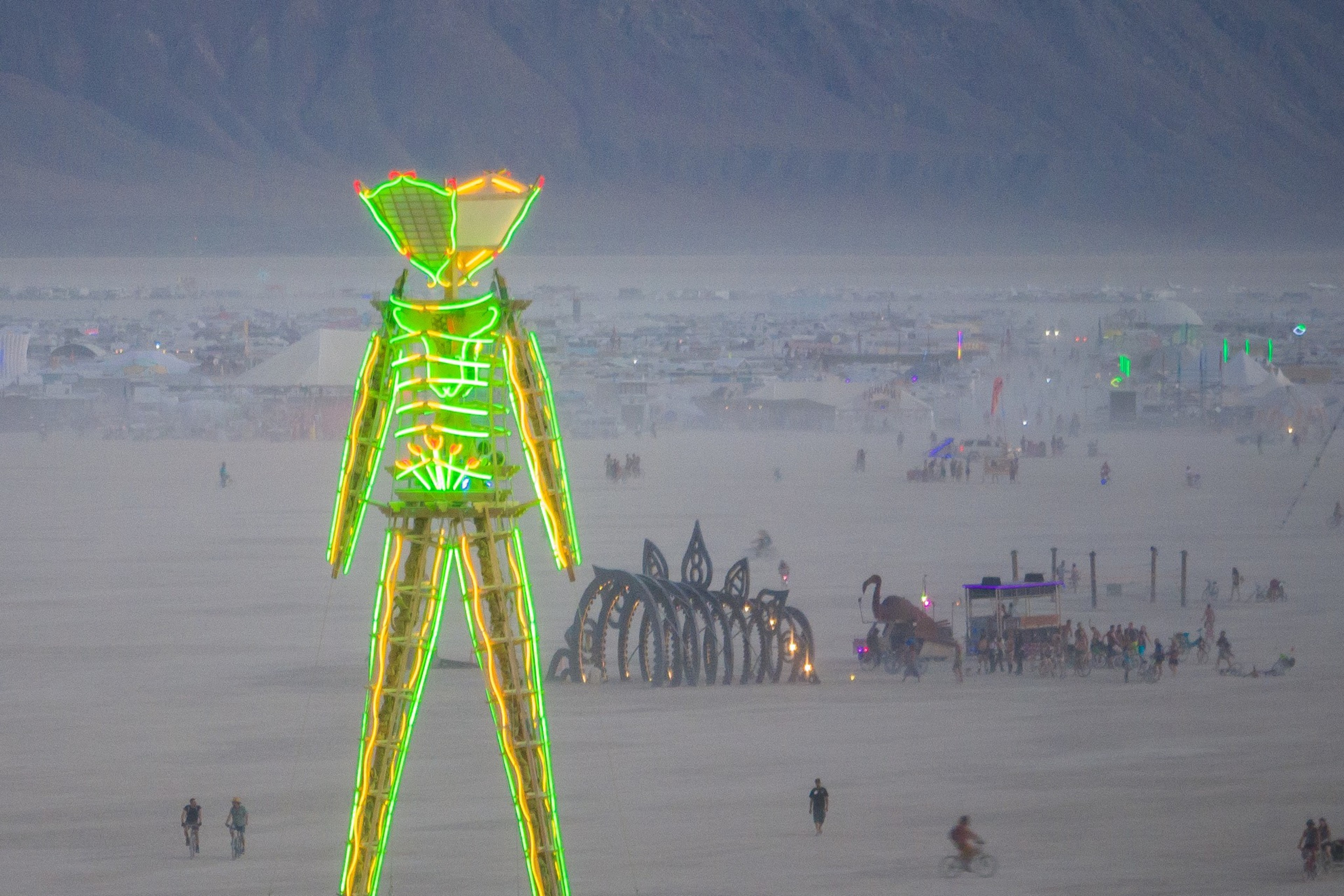 A tall, neon-lit structure resembling a person stands in a desert setting, with distant tents and art installations. People and bikes are scattered around the area.