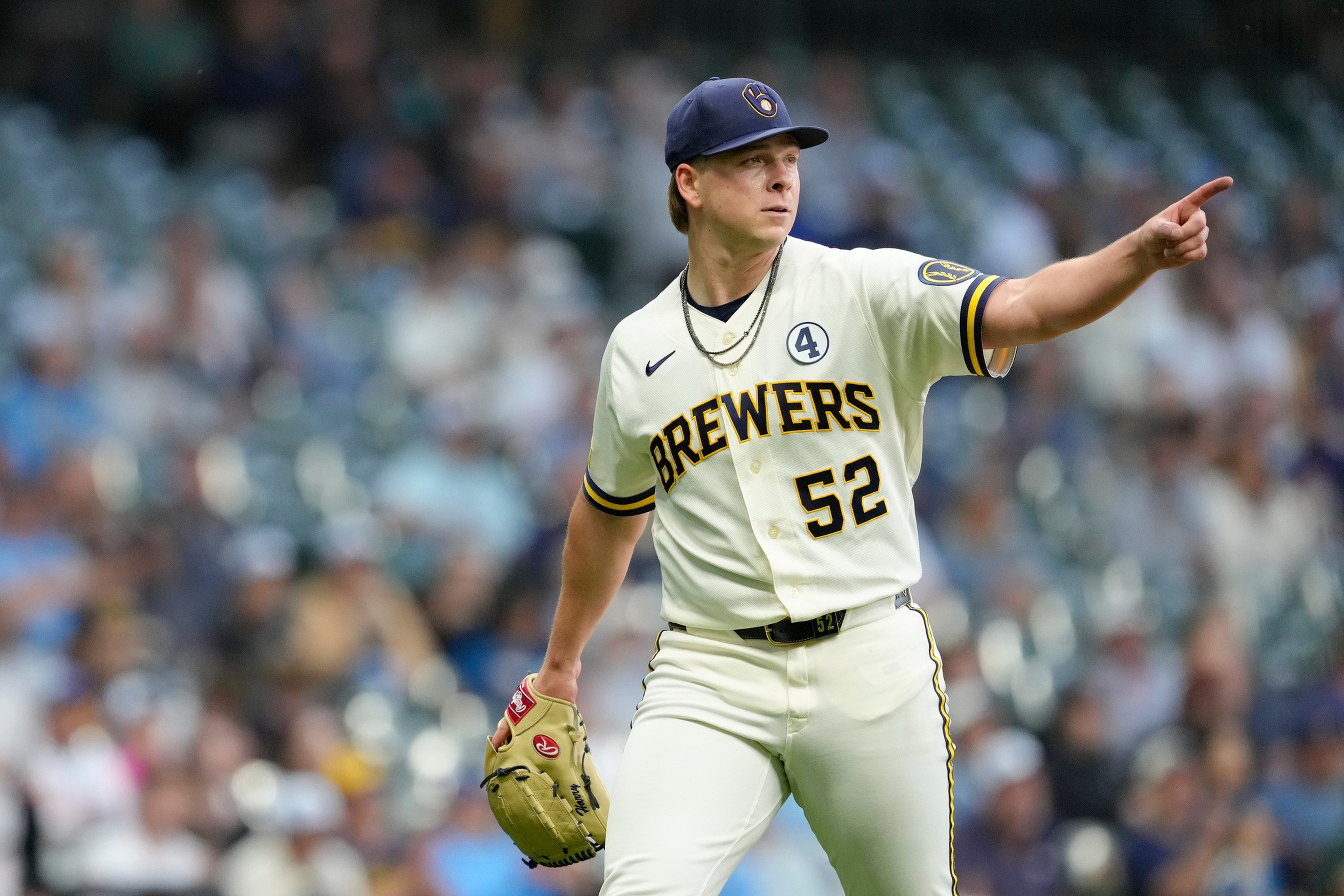 A baseball player in a Brewers uniform with number 52 is pointing with his right hand while holding a glove in his left hand on the field.