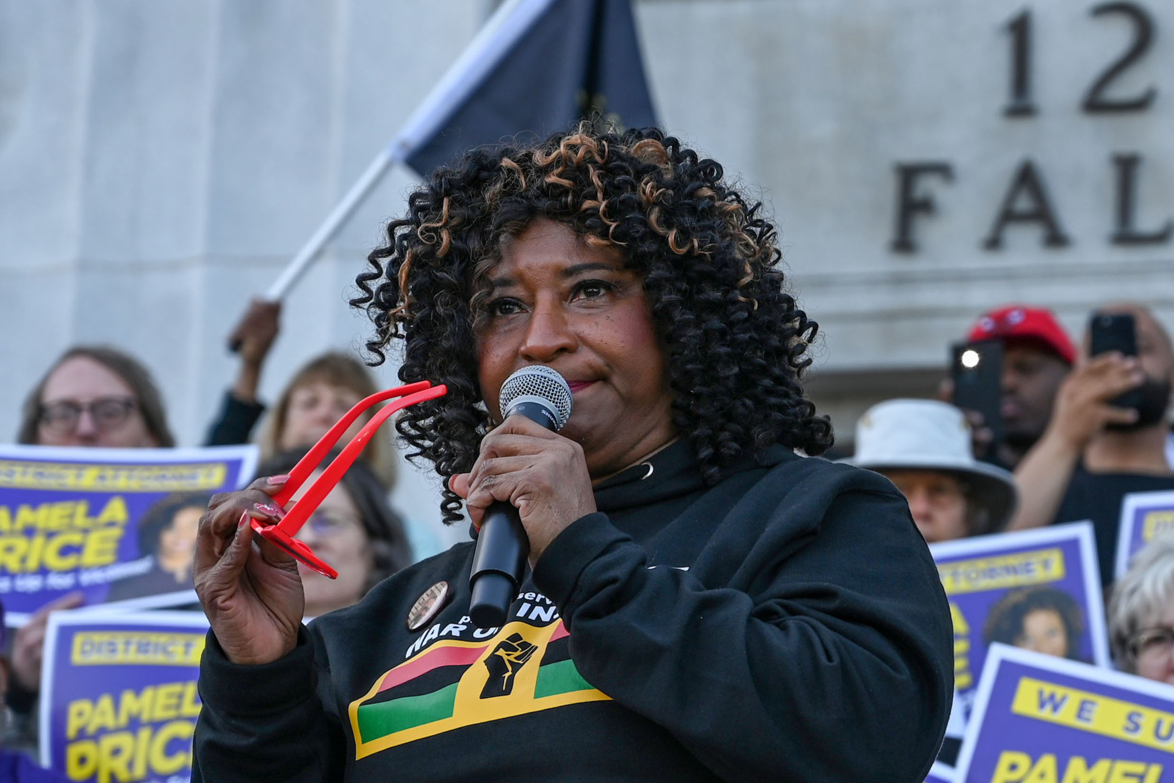 A woman with curly hair speaks into a microphone while holding red glasses, surrounded by people holding signs supporting Pamela Price.