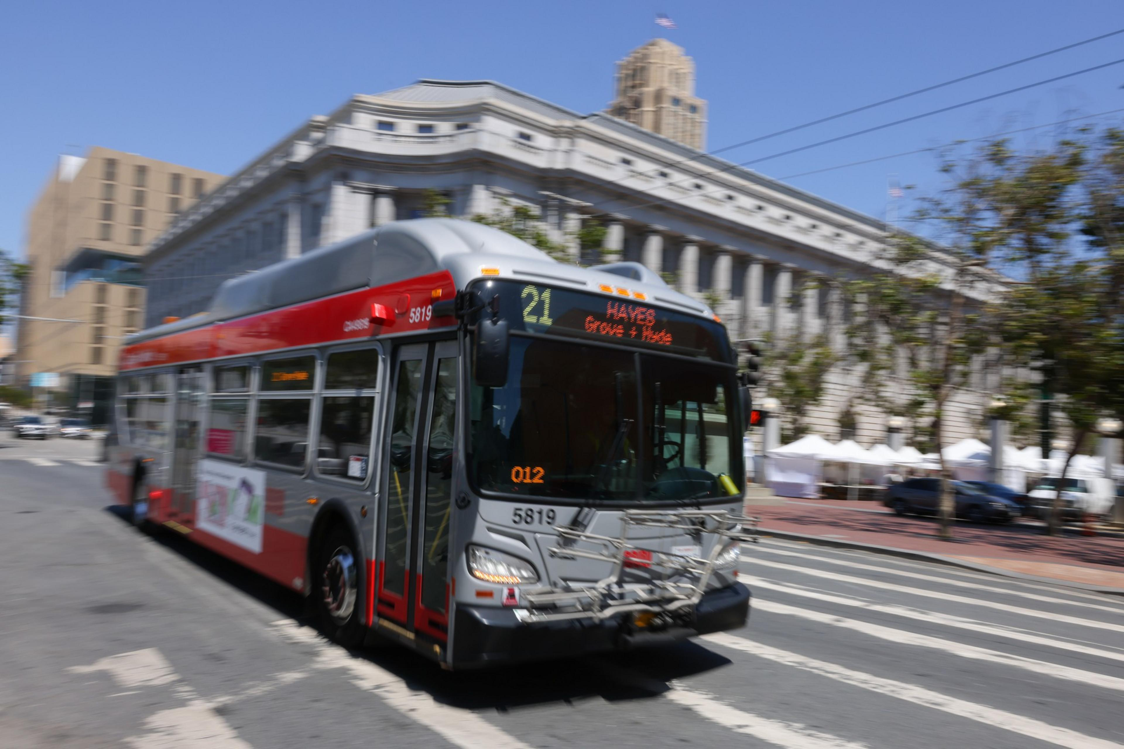 A gray and red city bus, marked route 21 to Hayes, moves along a crosswalk. It passes a large, classical-style building on a sunny day.