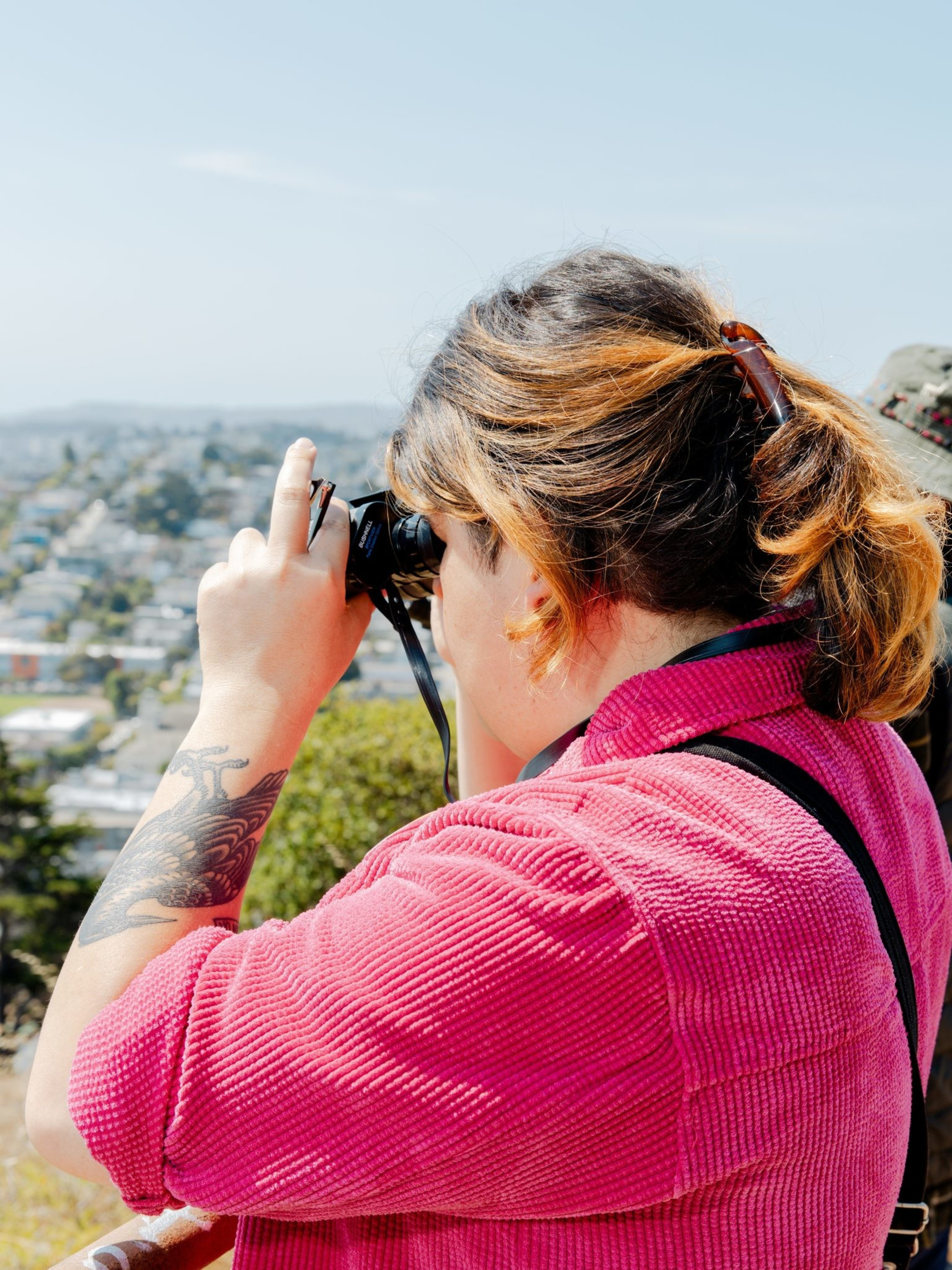 A person with a tattooed arm, wearing a pink jacket and sunglasses on their head, is taking a photo against a blurry urban landscape.