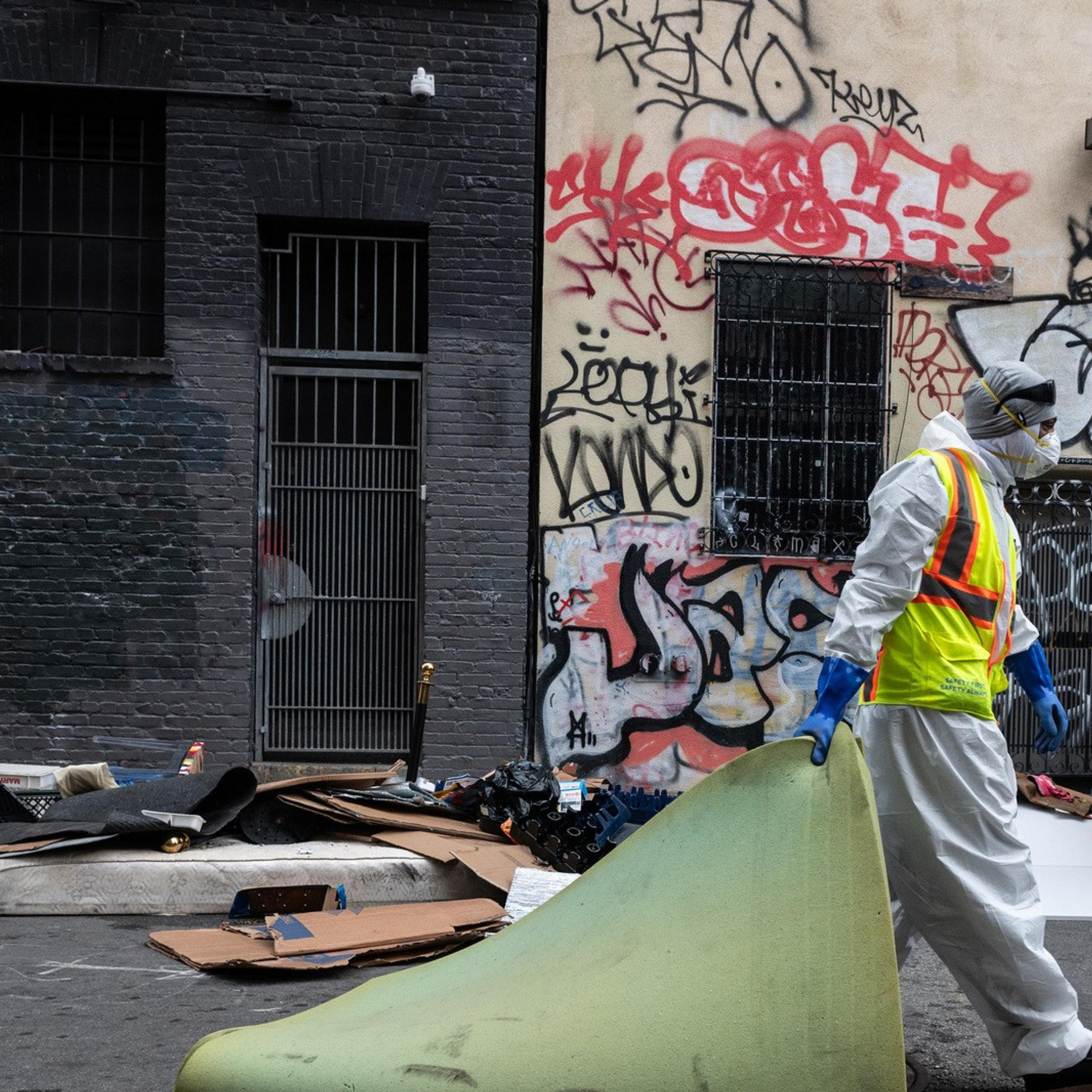 A person in protective gear carries a rolled-up carpet in an alley with graffiti-covered walls. Debris and a truck labeled "DPW" are visible around.