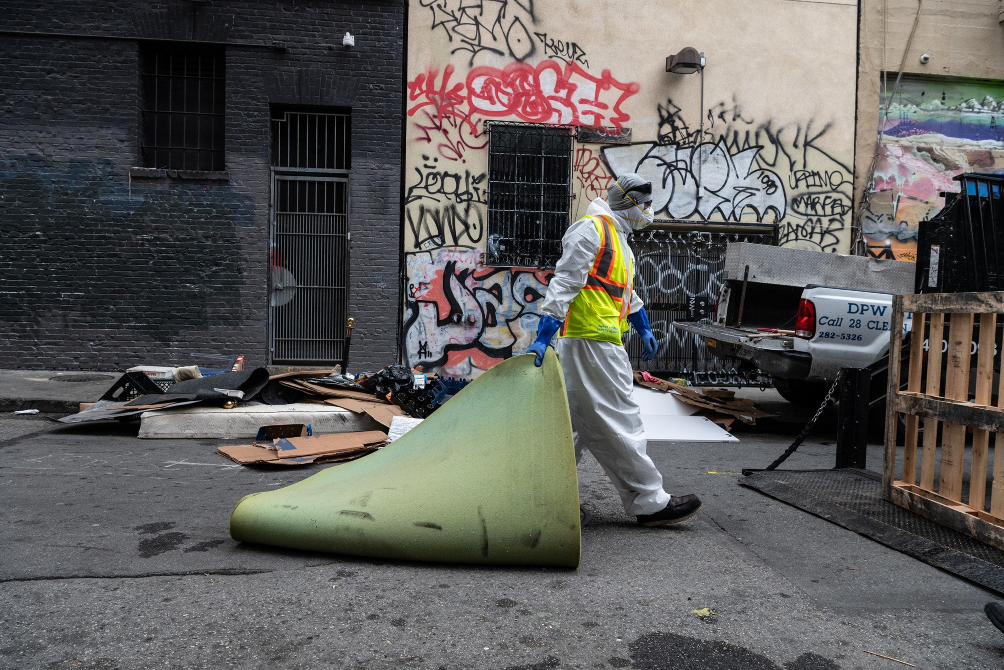 A person in protective gear carries a rolled-up carpet in an alley with graffiti-covered walls. Debris and a truck labeled "DPW" are visible around.
