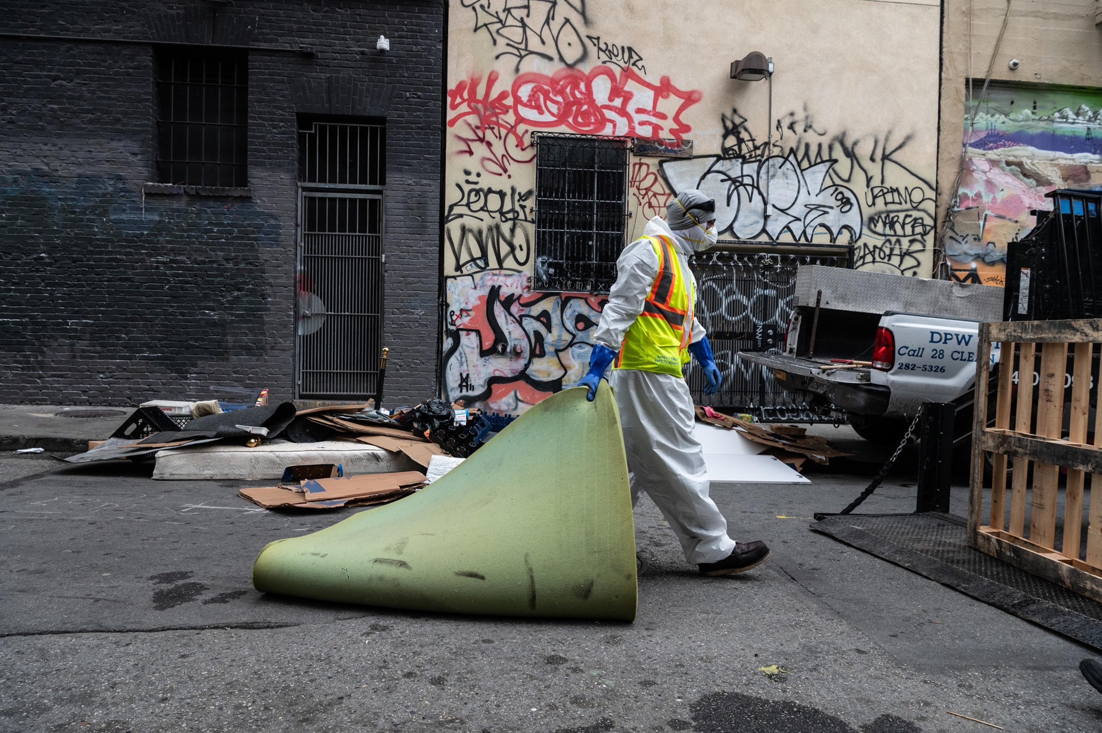 A person in protective gear carries a rolled-up carpet in an alley with graffiti-covered walls. Debris and a truck labeled "DPW" are visible around.
