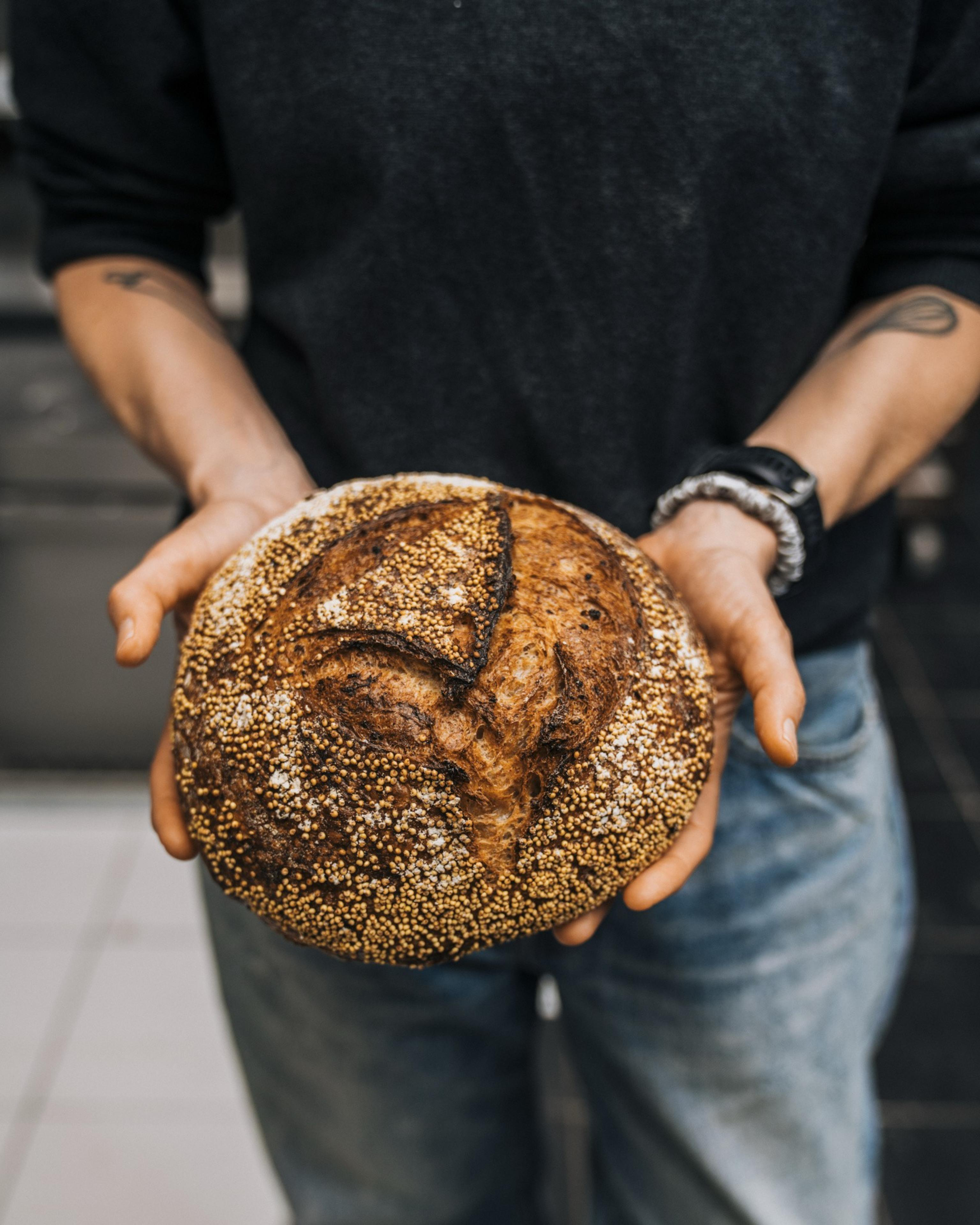 A person in a dark sweater holds a round, crusty loaf of bread sprinkled with seeds. The person is wearing jeans and a watch, with tattoos visible on their arms.