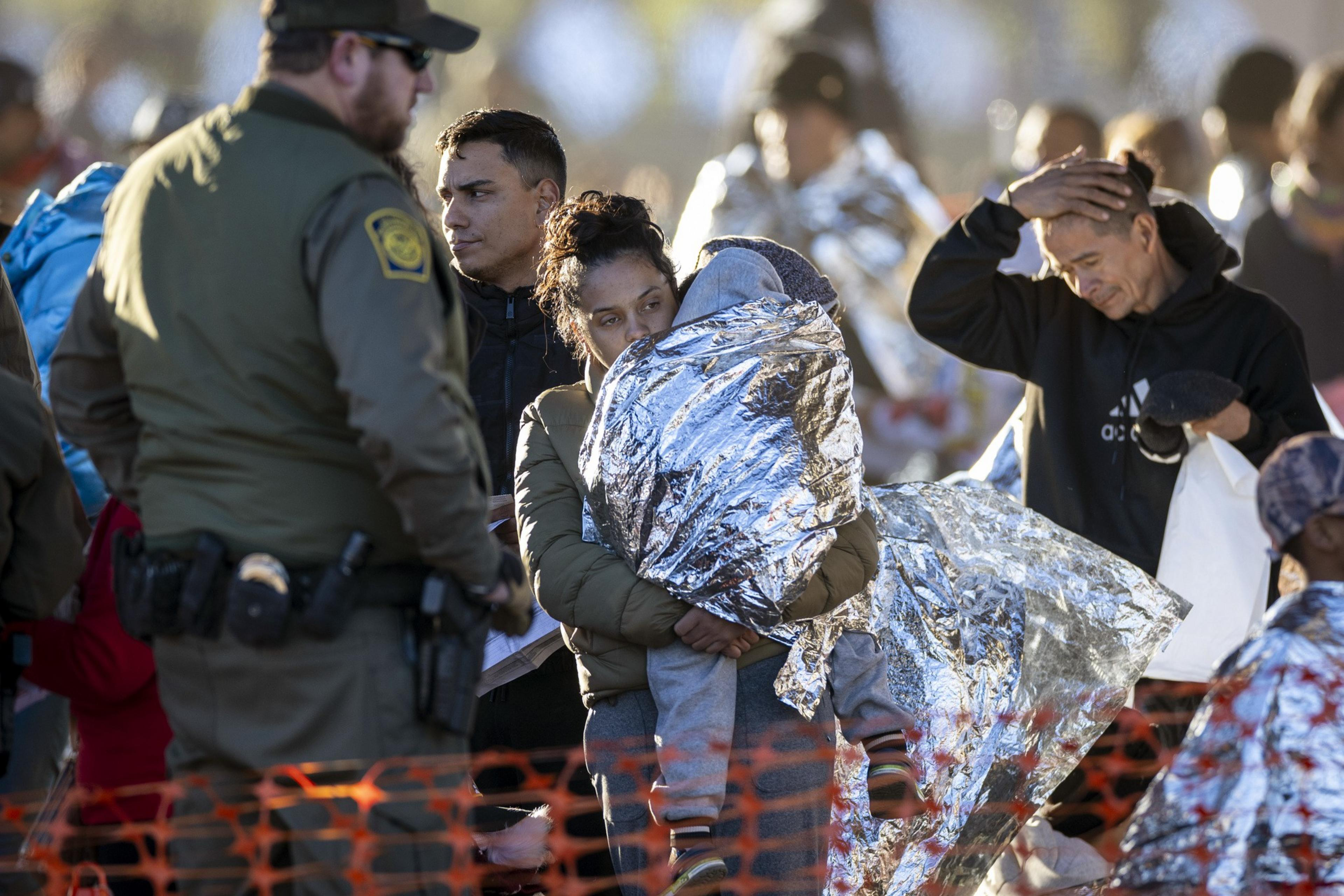 A woman holds a child wrapped in a reflective blanket, overseen by a border patrol officer. A man looks concerned.
