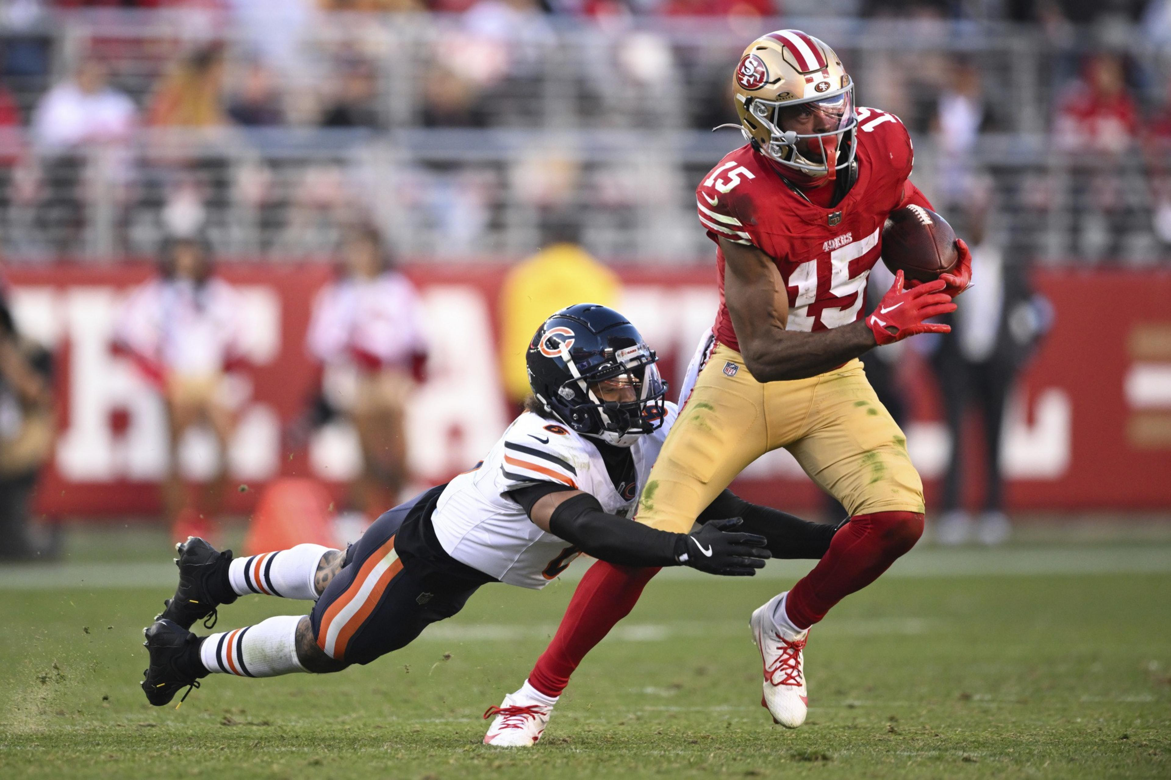 A football player in a red and yellow uniform runs with the ball while a player in a white and black uniform dives to tackle him.