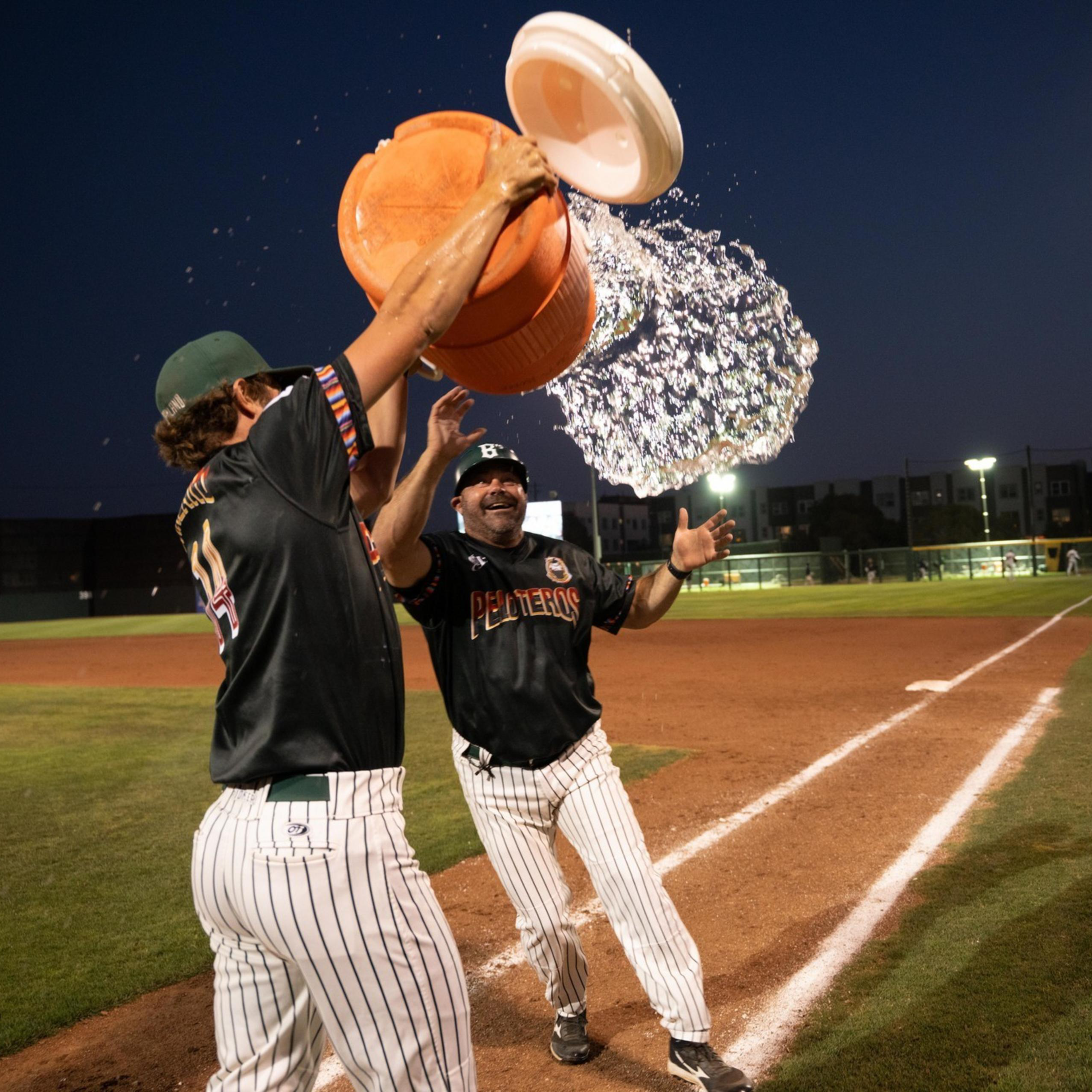 A baseball player is pouring a large container of water over a coach’s head on the field at night, celebrating with smiles under stadium lights.