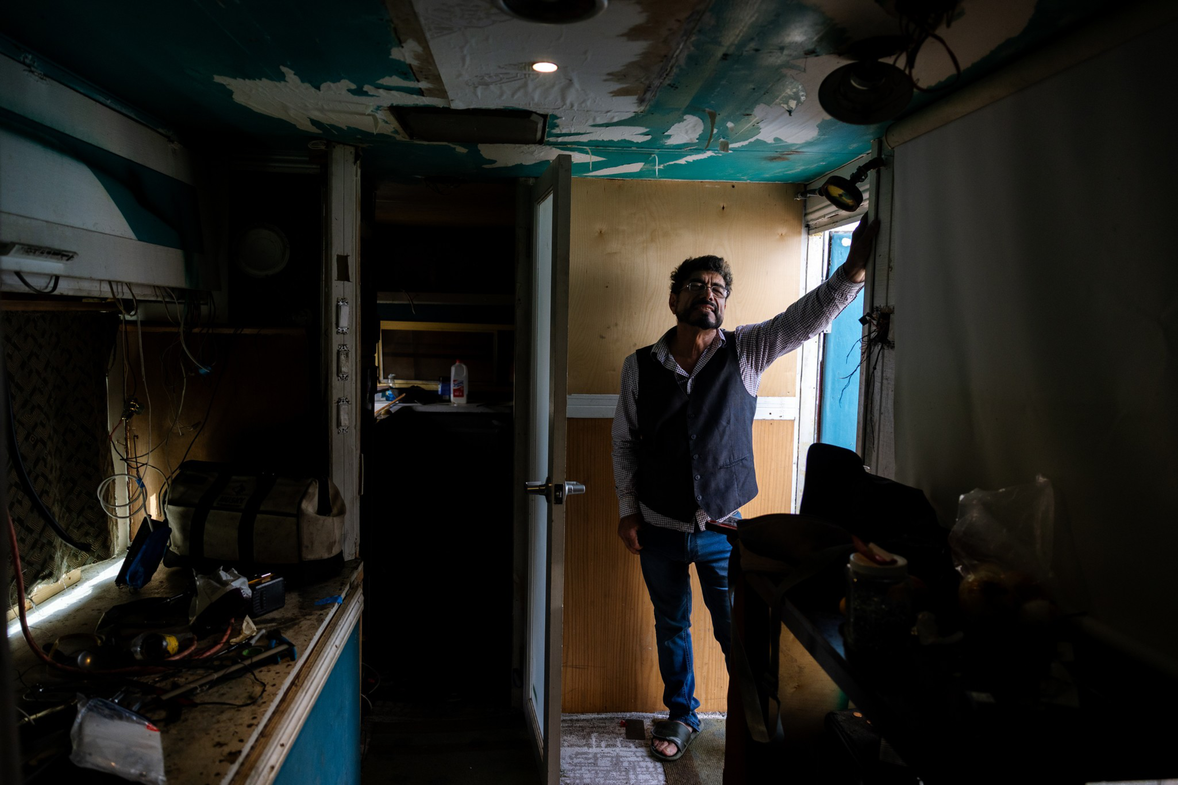 A man stands in a dimly lit, cluttered room with peeling paint, resting his hand on the wall near a small window with light streaming in.