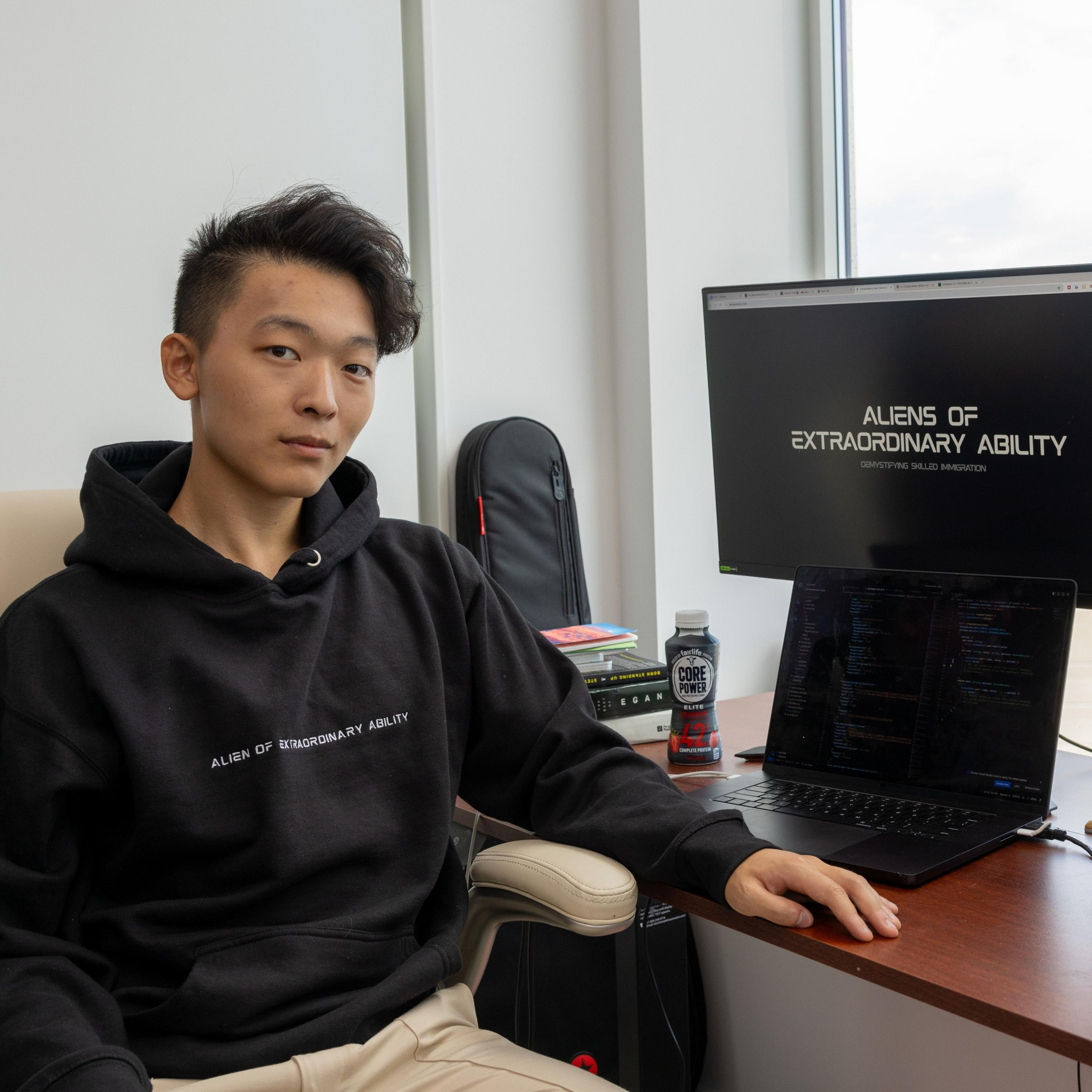 A person in a black hoodie sits at a desk with a laptop, a monitor, books, and a drink. The monitor displays &quot;ALIENS OF EXTRAORDINARY ABILITY.&quot;