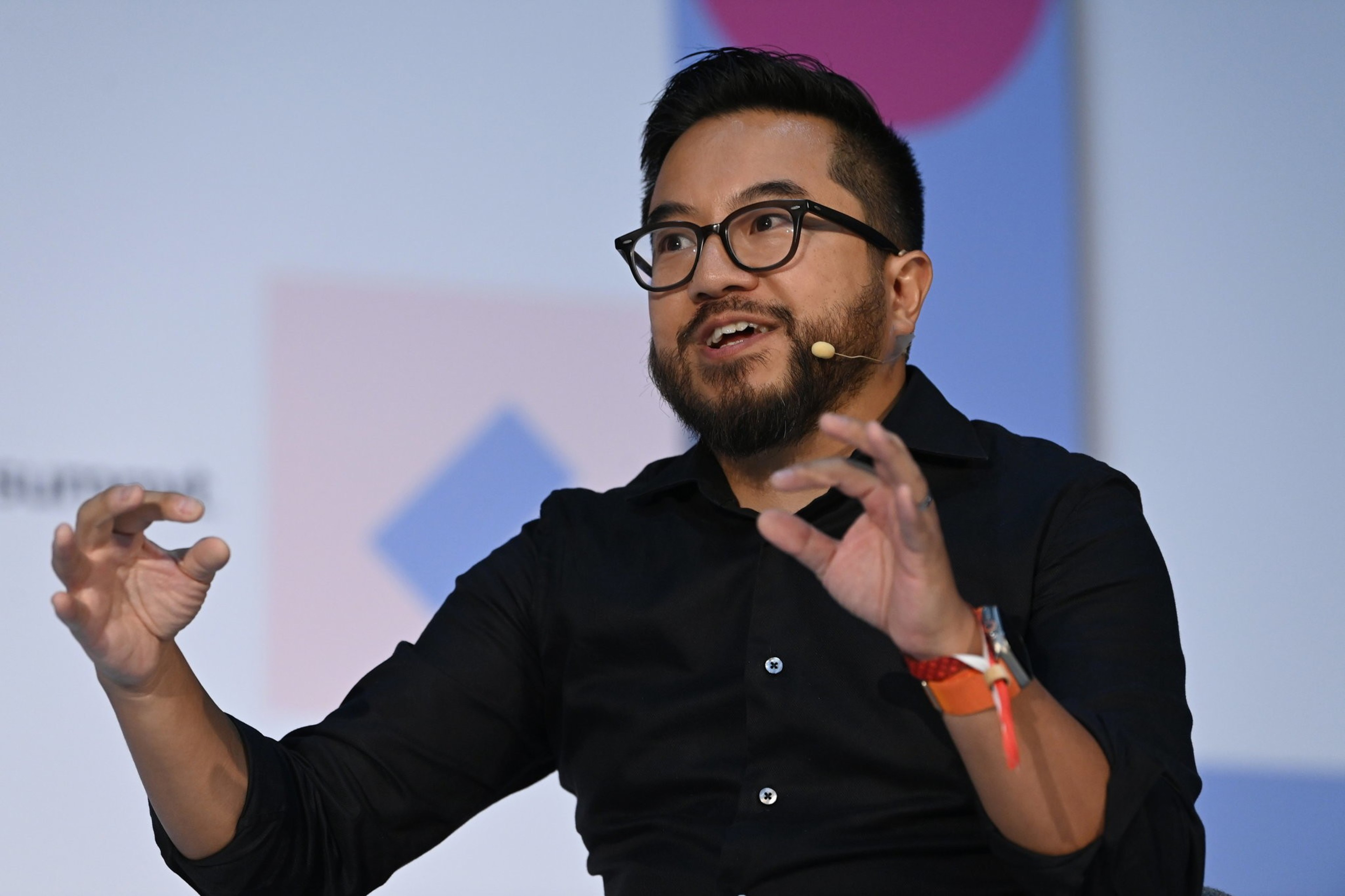 A man with glasses and a beard is speaking animatedly into a headset microphone. He's wearing a black shirt and gesturing with his hands during a presentation.