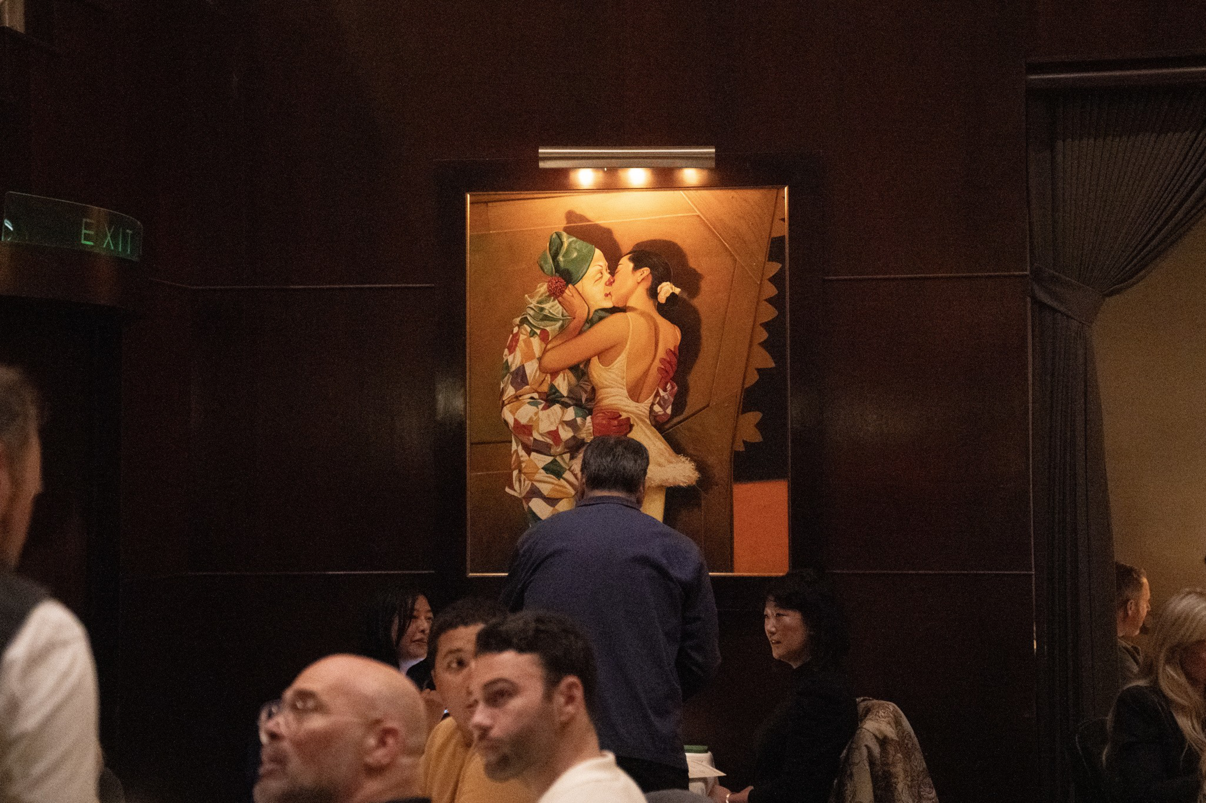 A man stands closely facing a large painting of two women embracing and kissing, surrounded by seated restaurant guests in a dimly lit room.