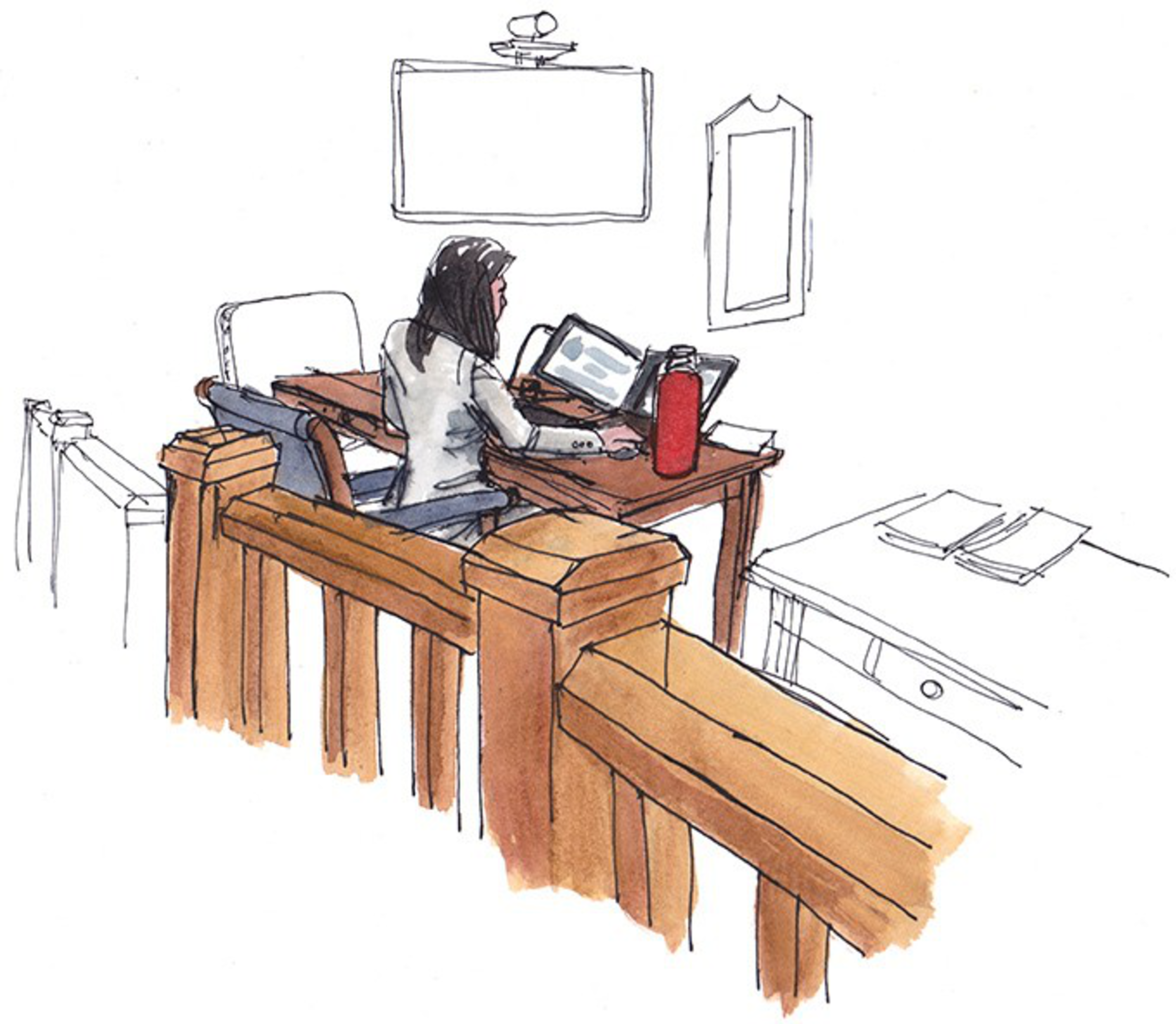 A woman sits at a wooden desk working on a laptop, with a red water bottle nearby, a wooden railing in the foreground, and papers on a nearby table.