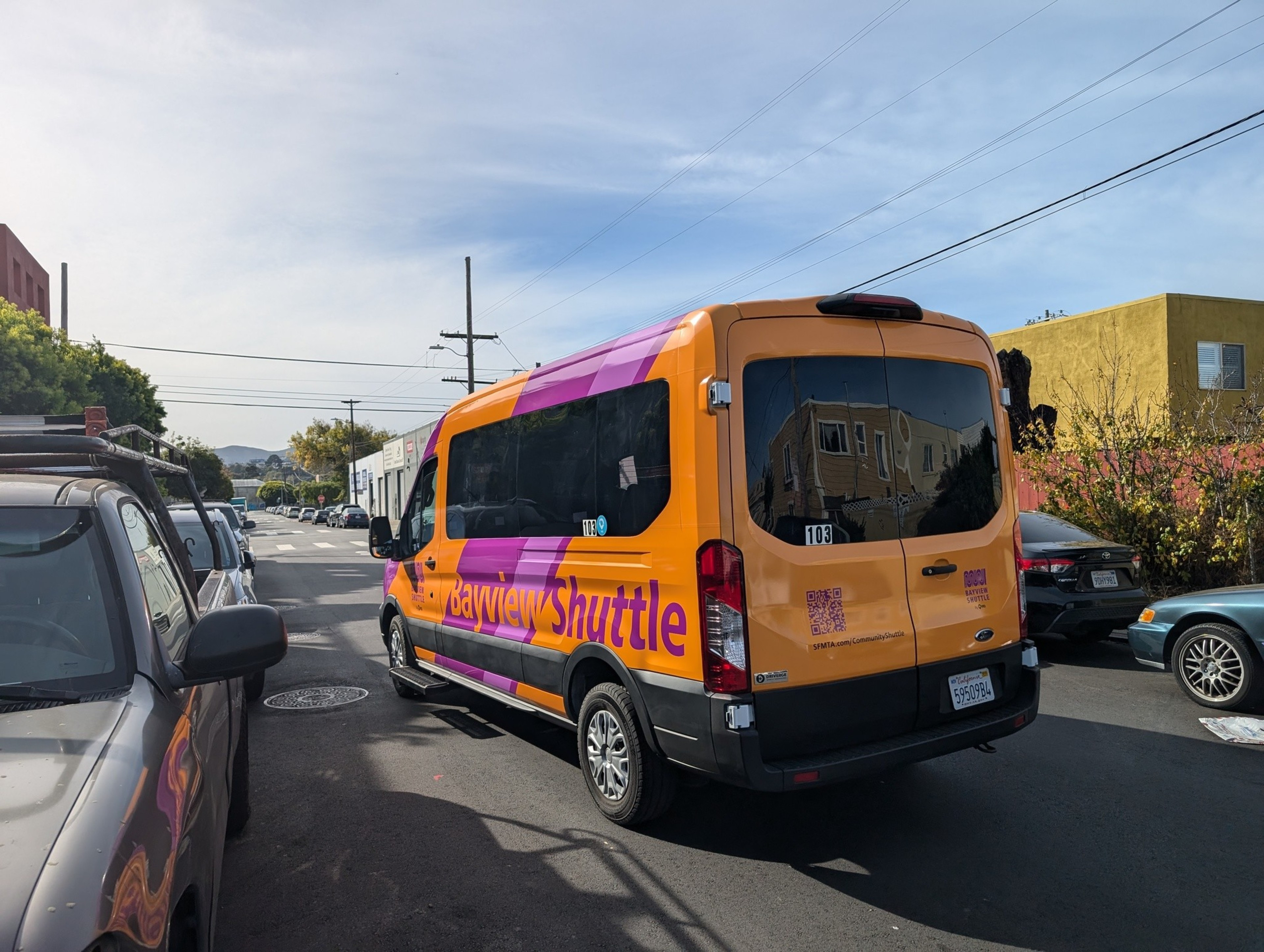 A yellow and purple Bayview Shuttle van is parked on a sunny street, surrounded by other parked cars and some buildings in the background.