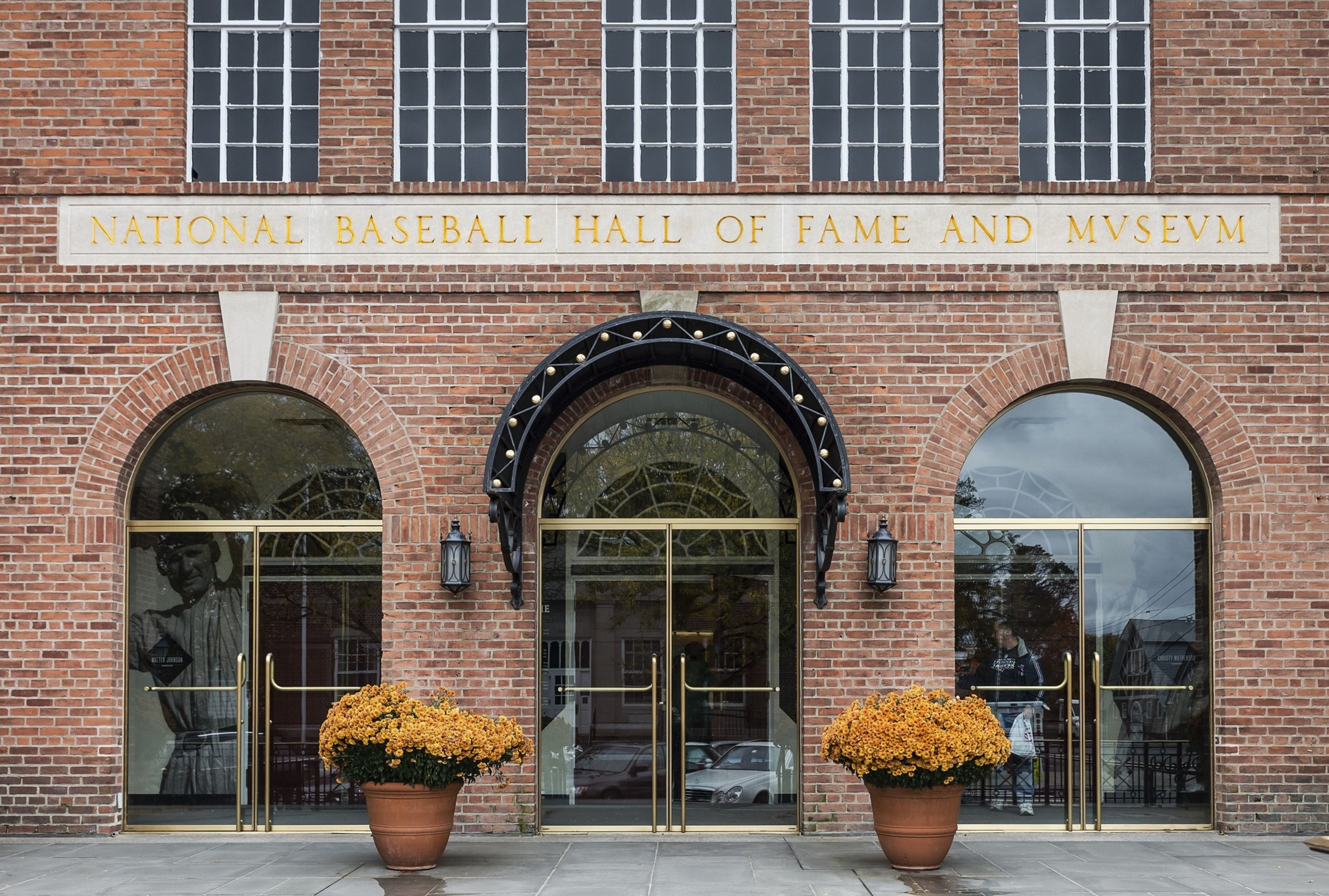 A brick building facade with three arched glass doors, two potted orange flowers, and a sign reading "National Baseball Hall of Fame and Museum."