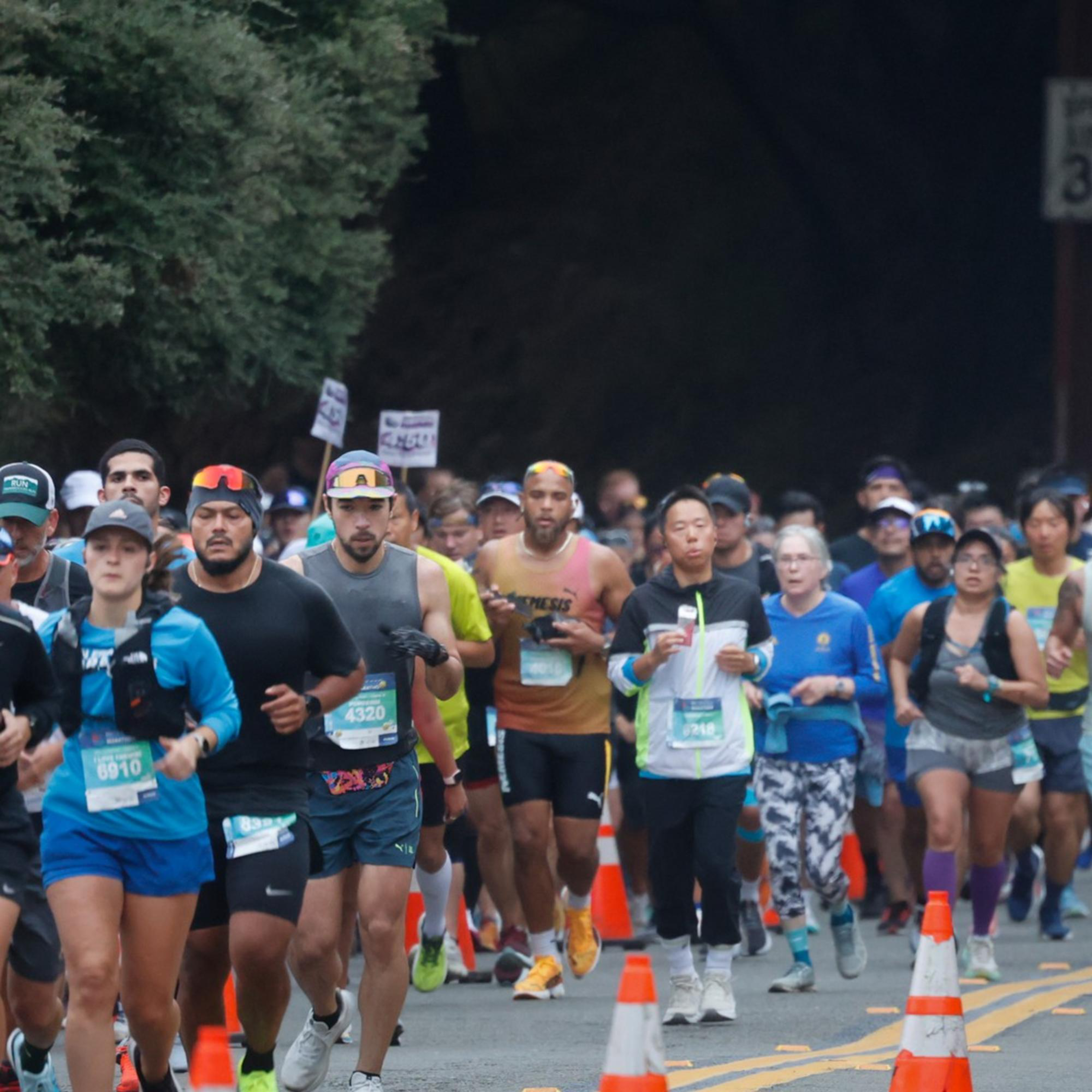 A group of runners in athletic gear is participating in a race on a road with traffic cones, while a motorbike rider and car are visible to the right.
