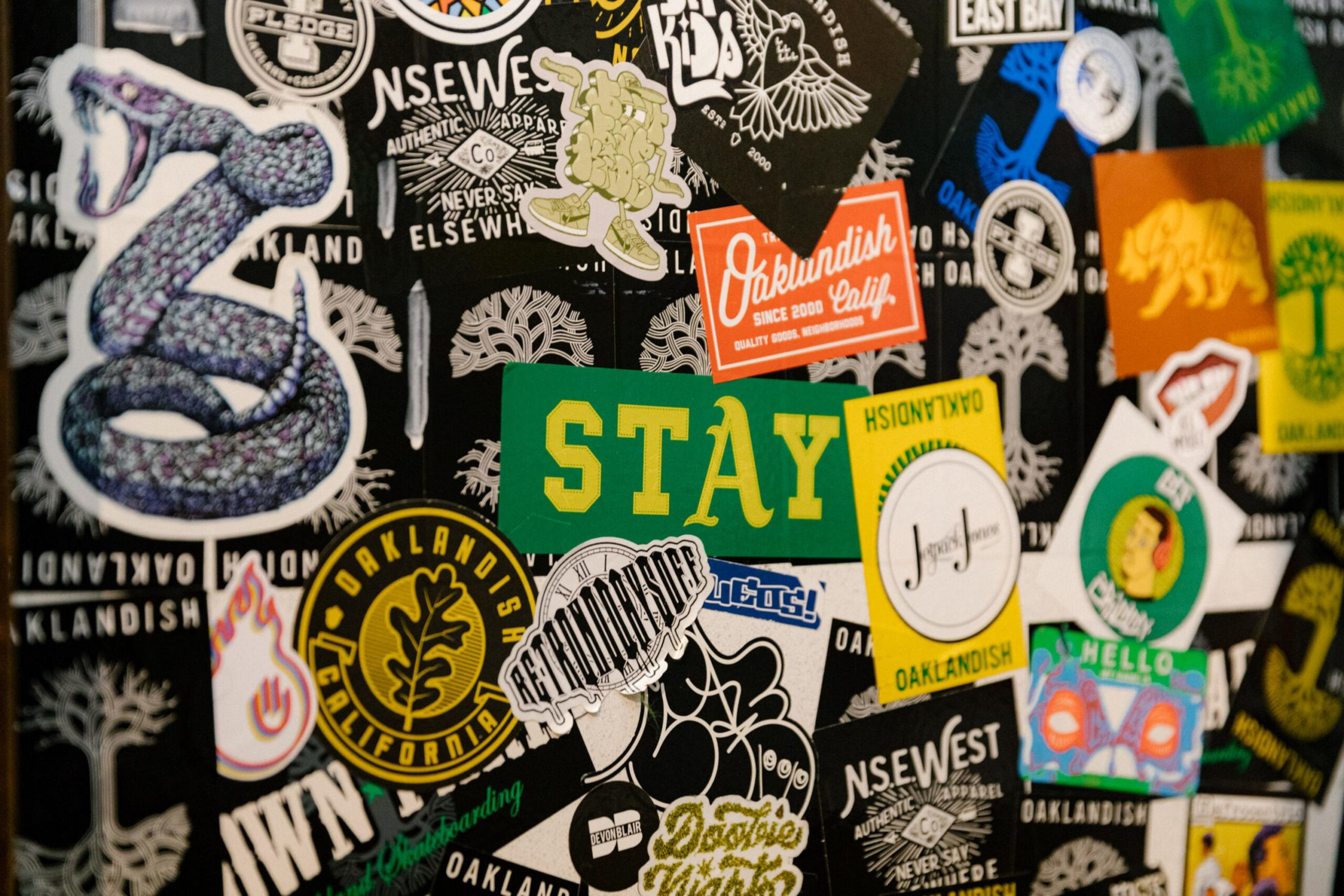 Various colorful stickers, including a green "STAY" sticker, a purple snake, and several Oakland-themed designs, are arranged on a black surface.