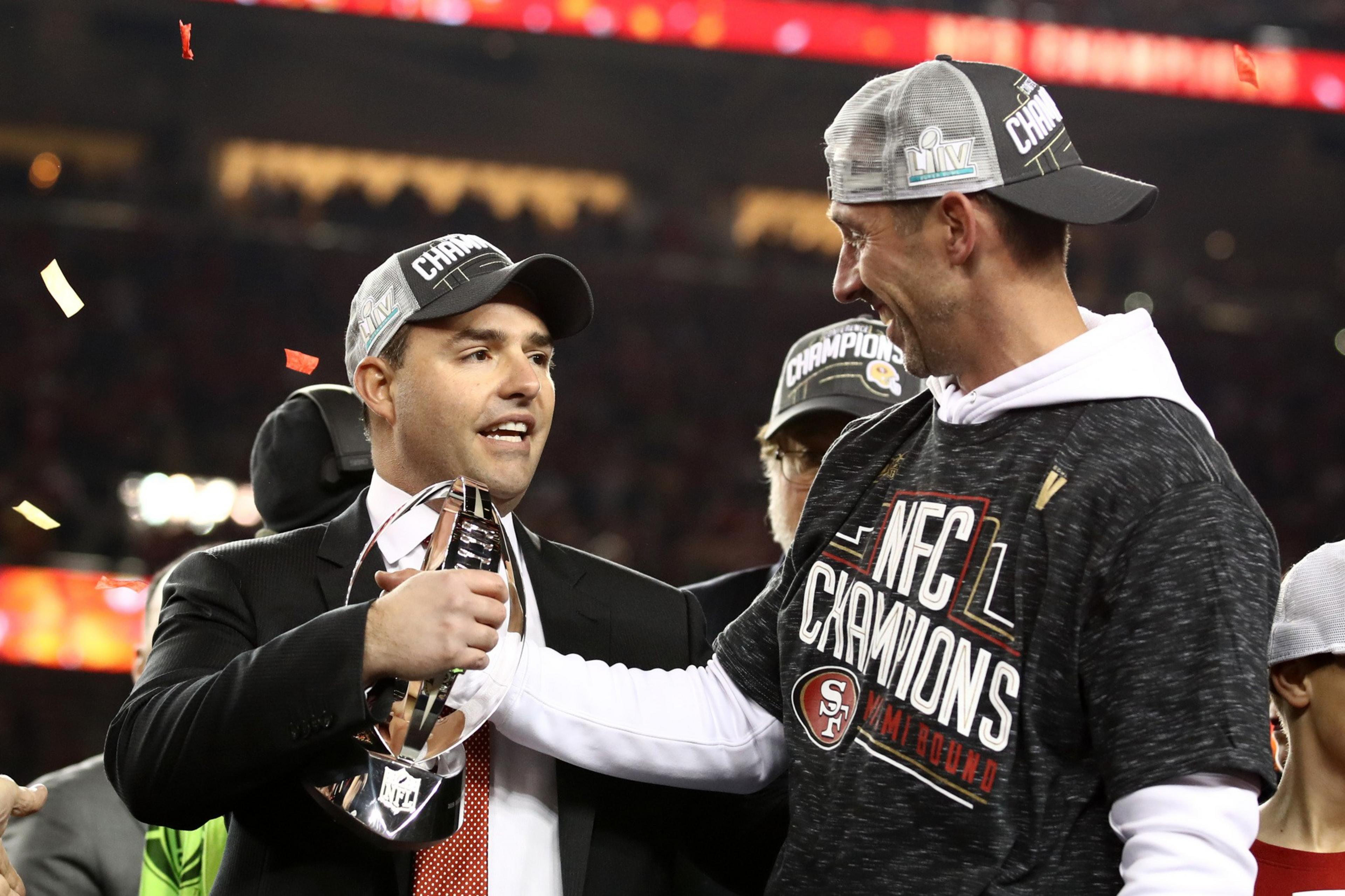 Two men wearing &quot;Champions&quot; caps celebrate with a trophy amid falling confetti. One wears a suit, the other an &quot;NFC Champions&quot; shirt.