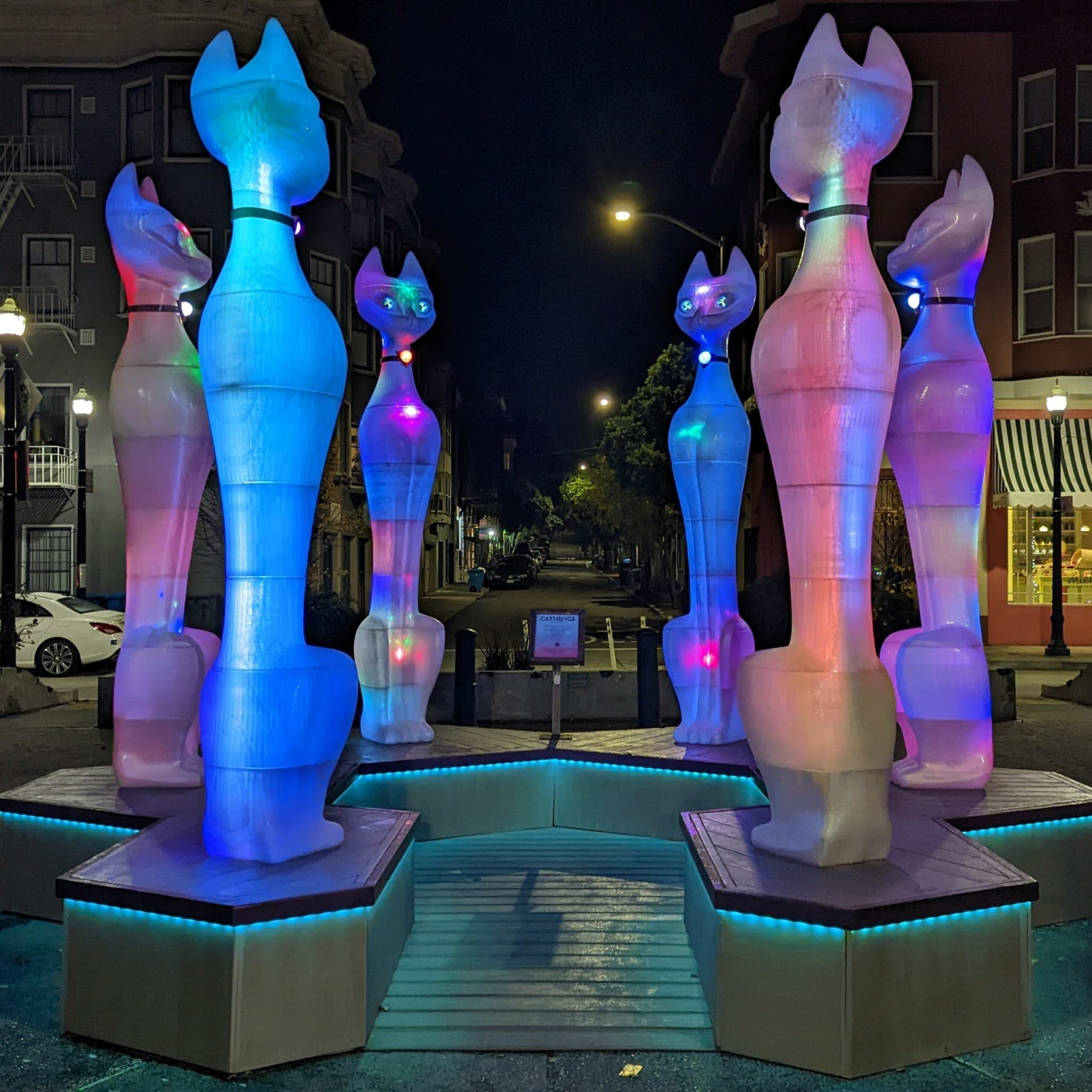 Six tall, glowing cat sculptures with colorful lights stand on platforms arranged in a hexagon on a street at night.