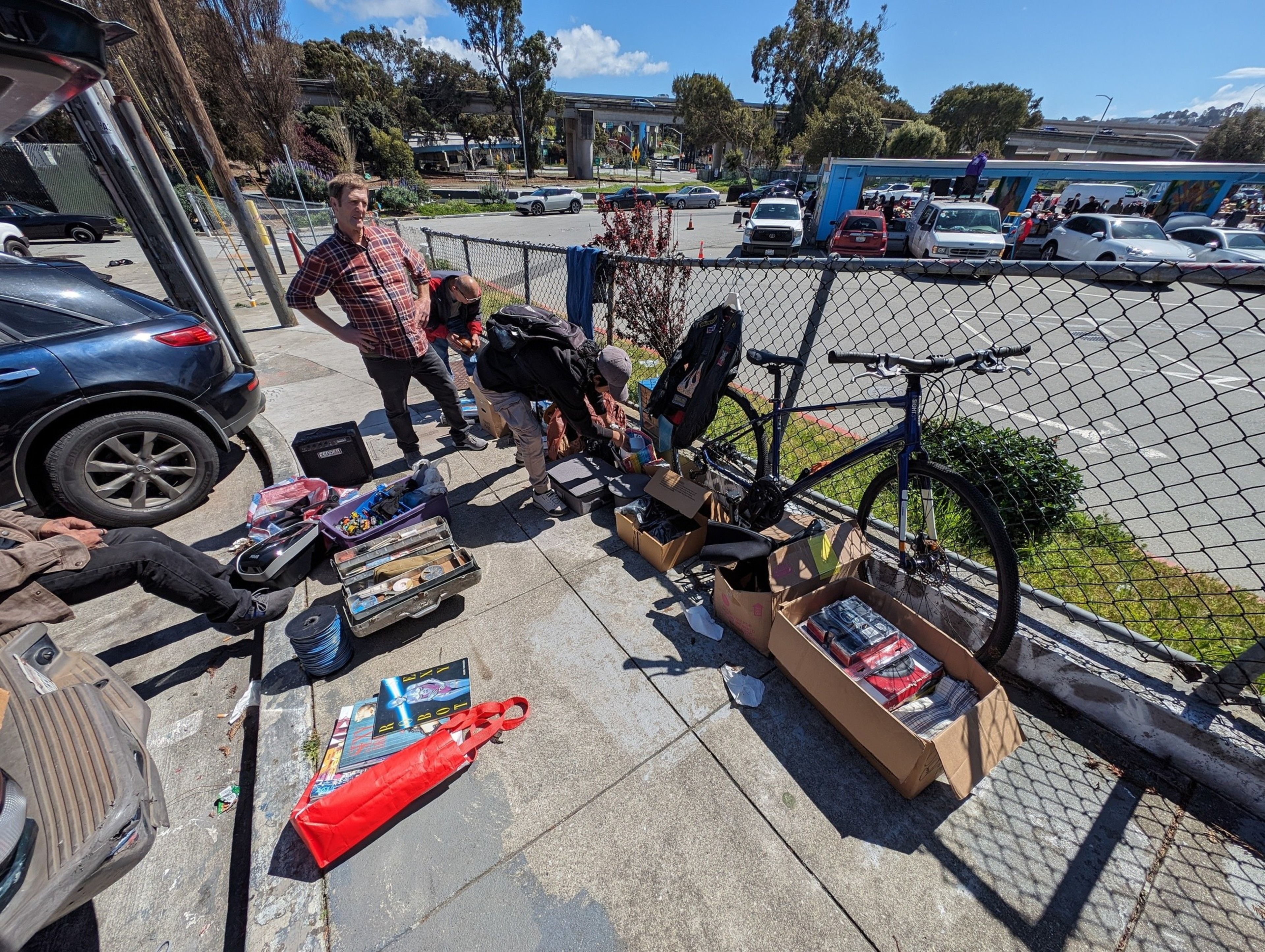 A man in a plaid shirt and jeans stands beside a row of items for sale along a sidewalk.