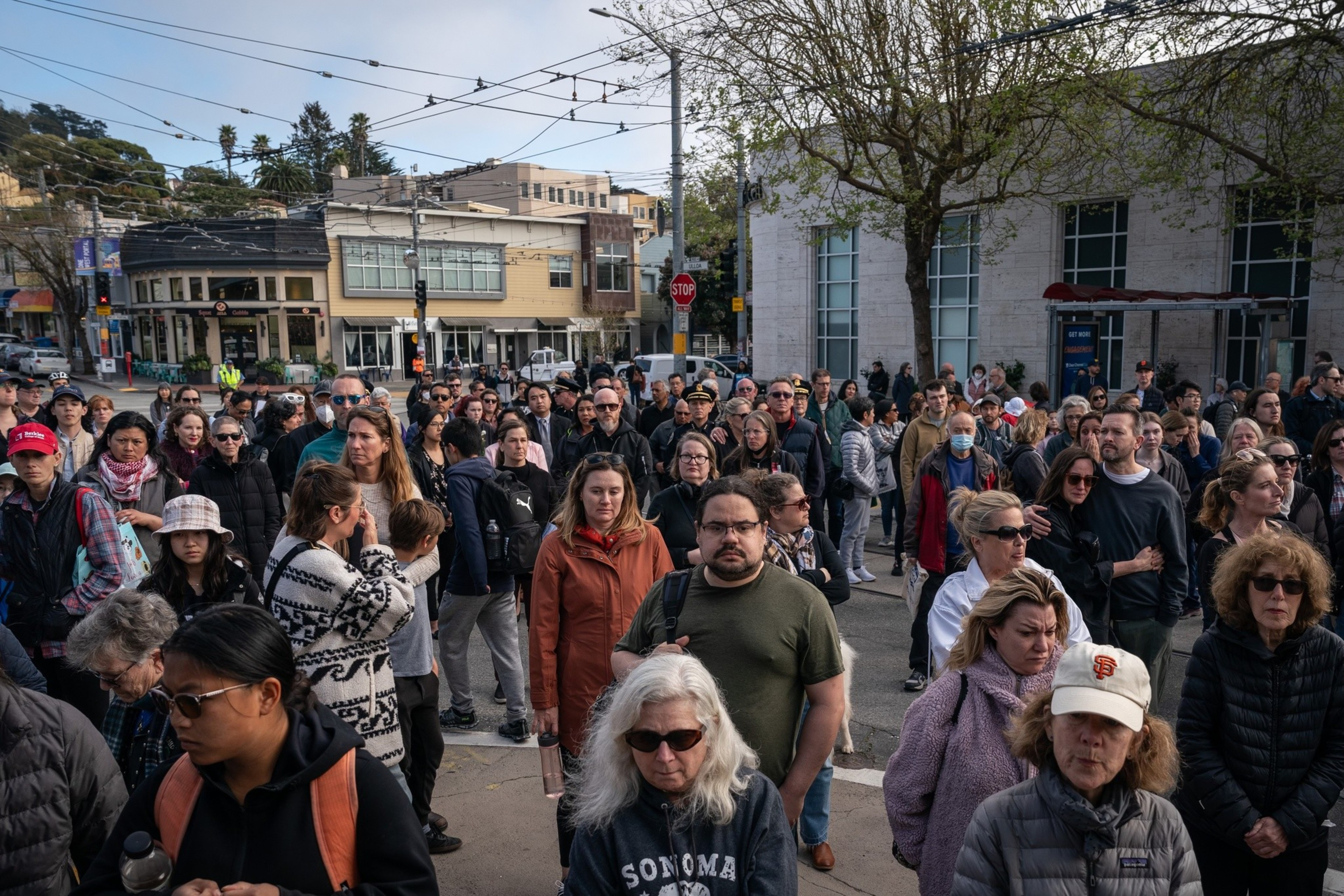 A crowded urban street with a diverse group of people, some wearing masks, under an overcast sky.