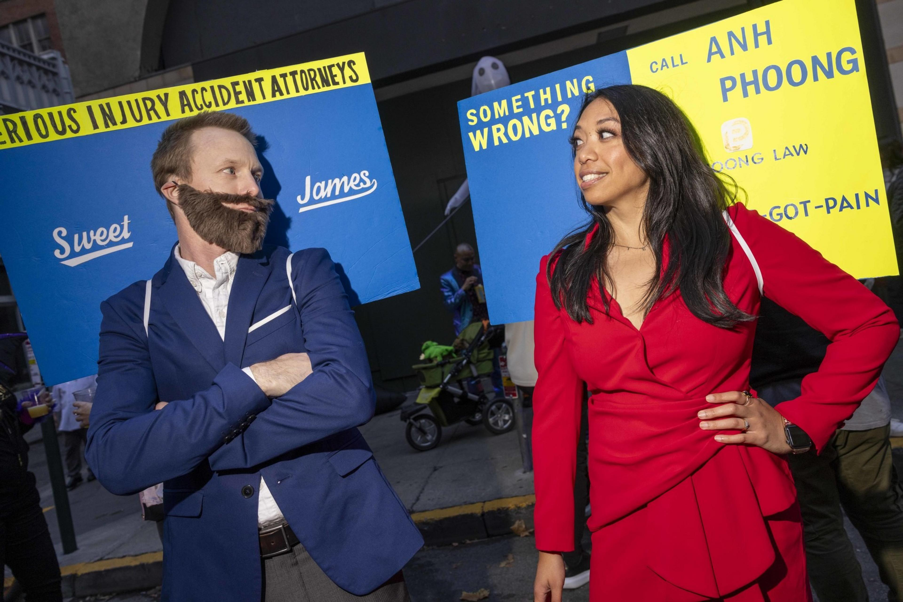 Two people in costumes stand facing each other with blue and yellow signs behind them, dressed as lawyers. One wears a blue suit, the other a red outfit.