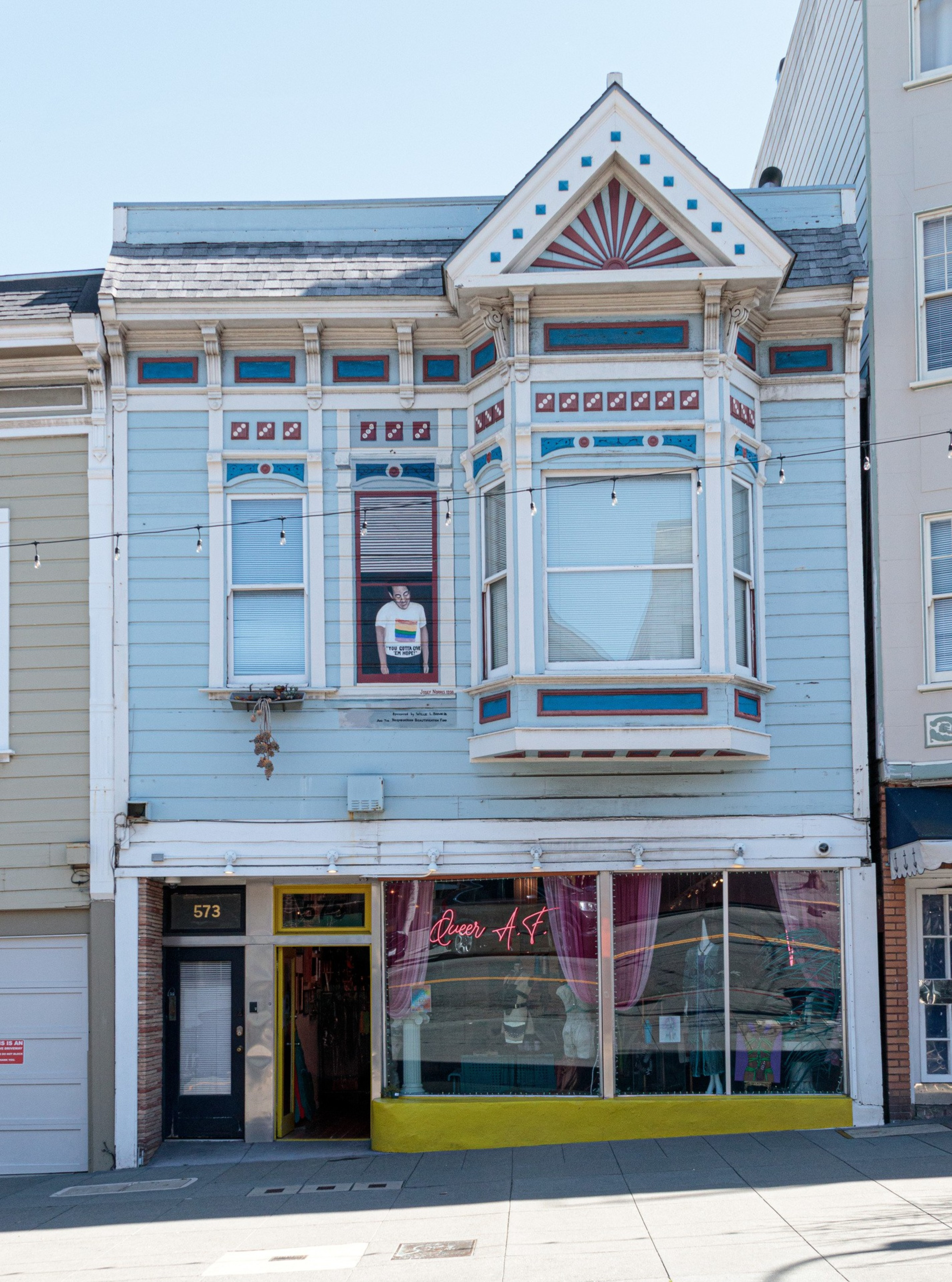 A two-story blue Victorian building with decorative trim has a mannequin wearing a white T-shirt in an upper window and a neon sign reading “Queer A.F.” in the ground floor display.