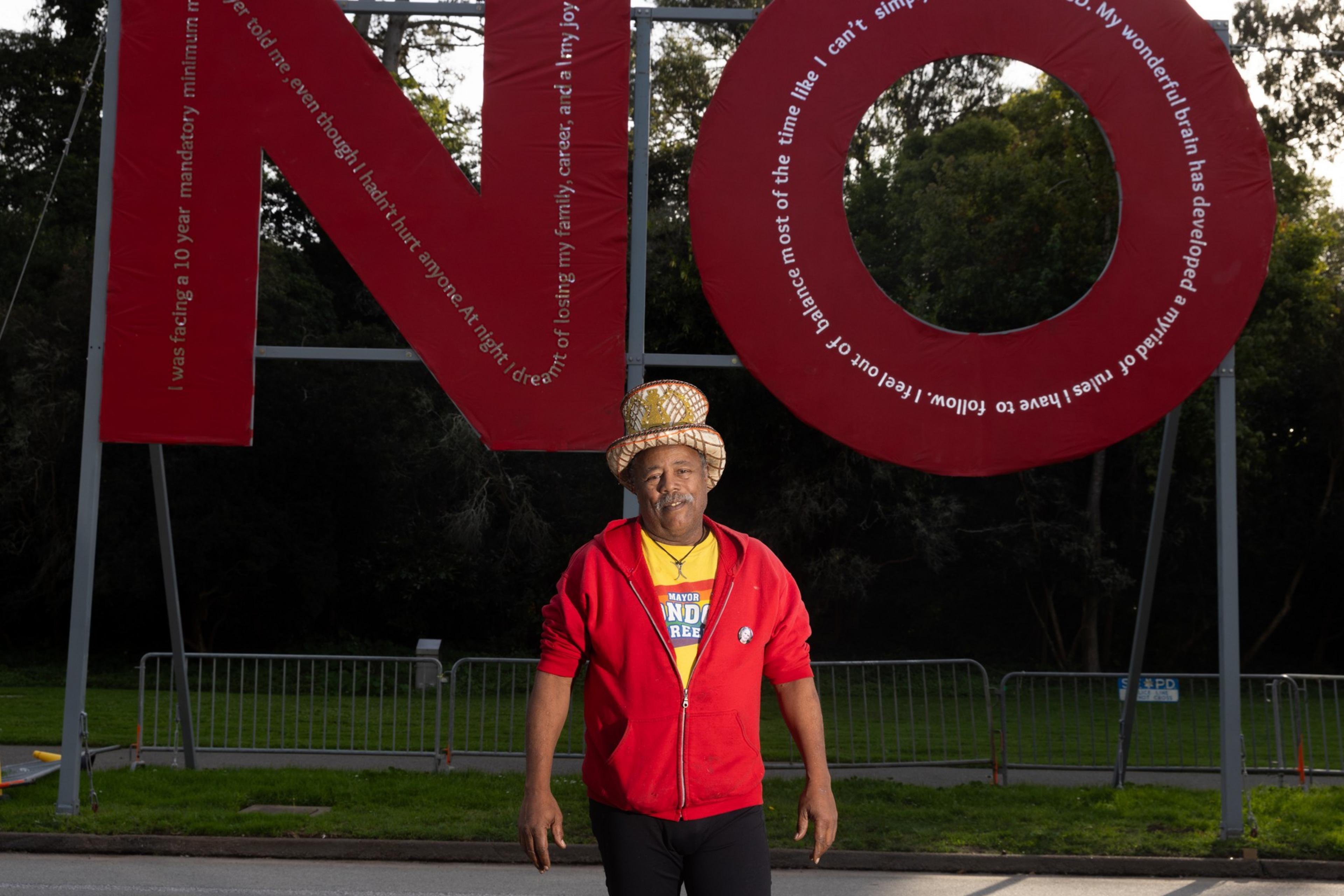 A man wearing a festive hat and red jacket stands in front of a large sculpture spelling "NO" with text around it, set against a backdrop of trees and fencing.