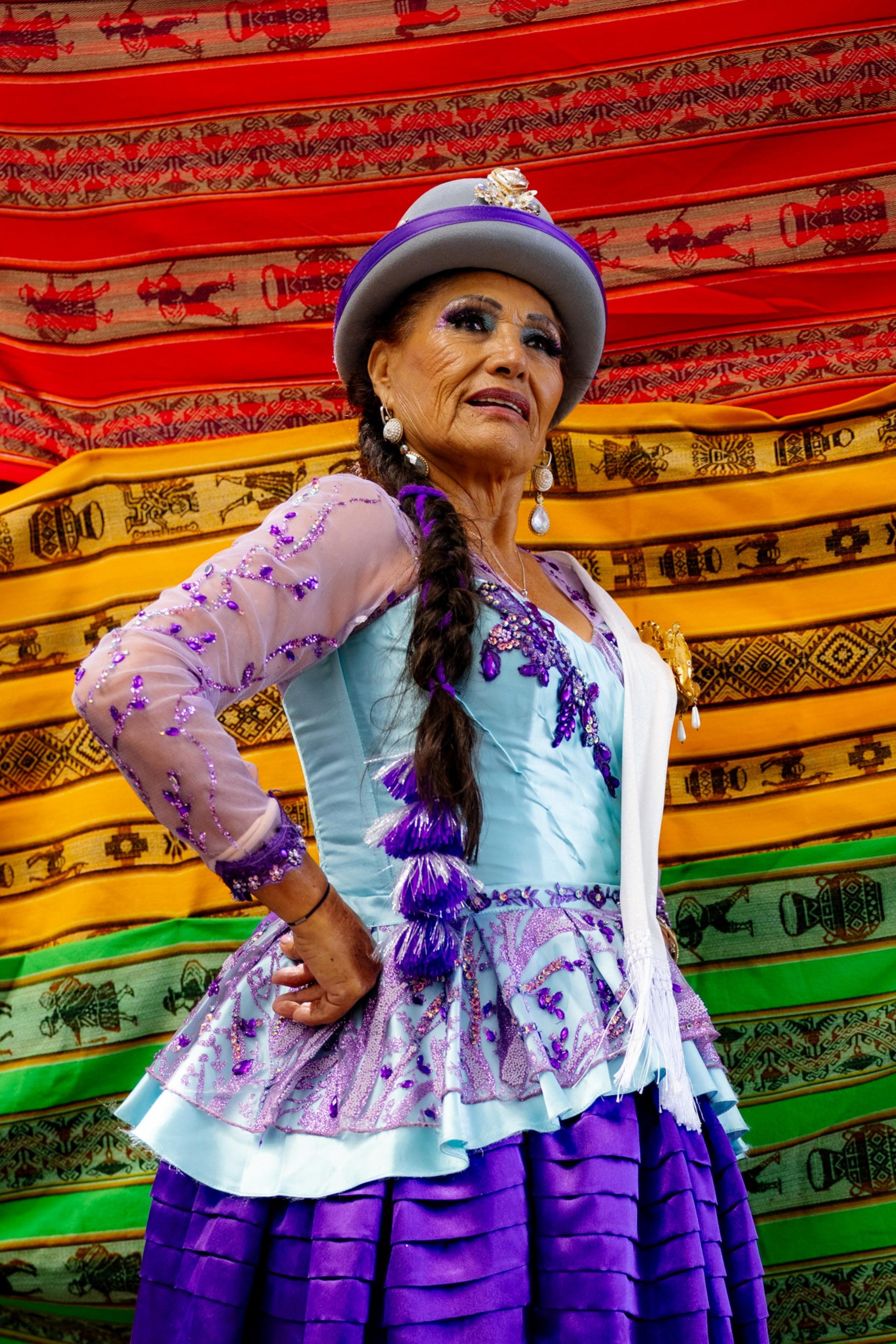 A woman in traditional attire stands confidently, wearing a colorful dress with purple sequins, a hat, and braided hair, against a vibrant, patterned backdrop.