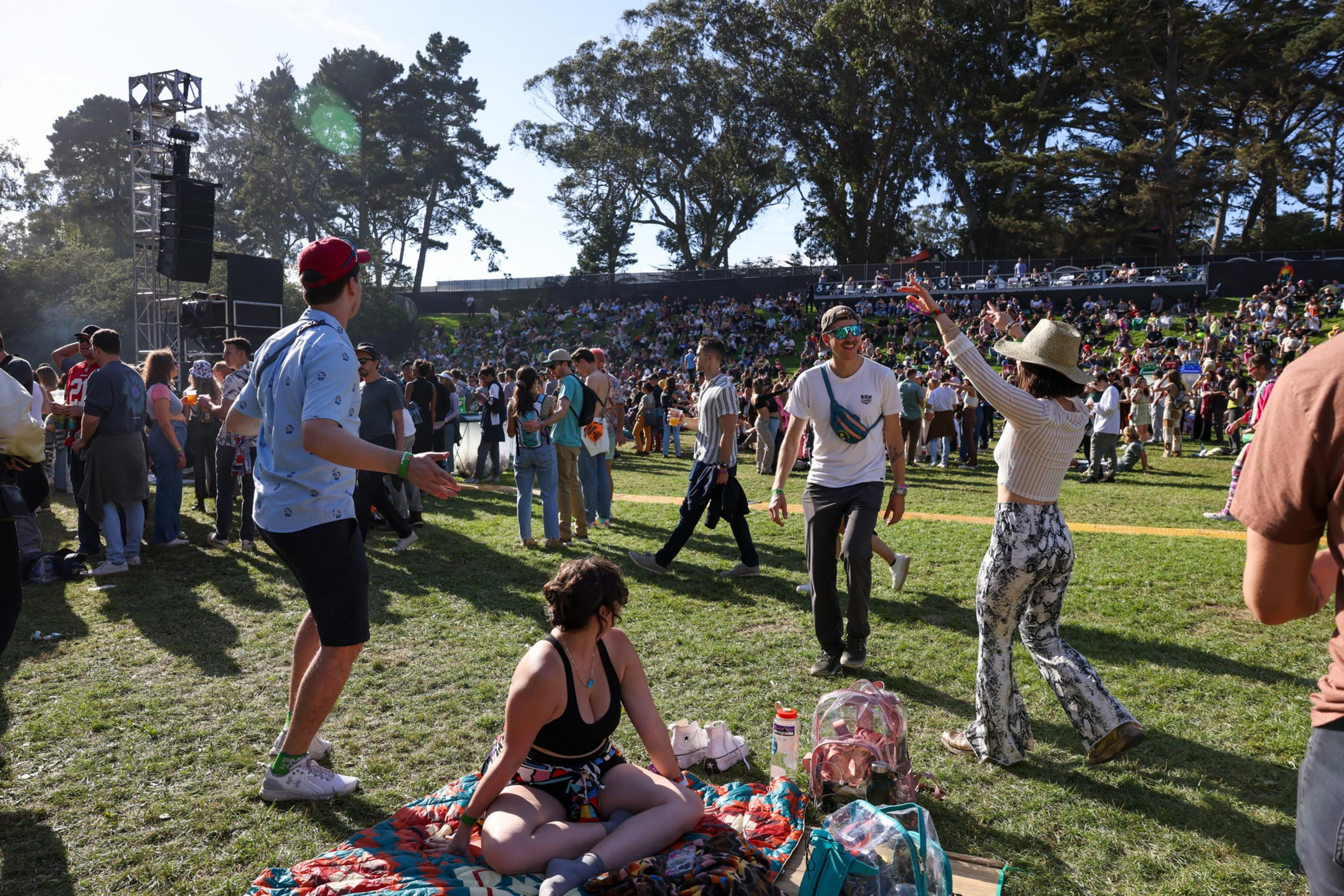 People enjoy an outdoor event on a grassy field with some dancing, sitting on blankets, and a large crowd in the background near trees and a speaker tower.
