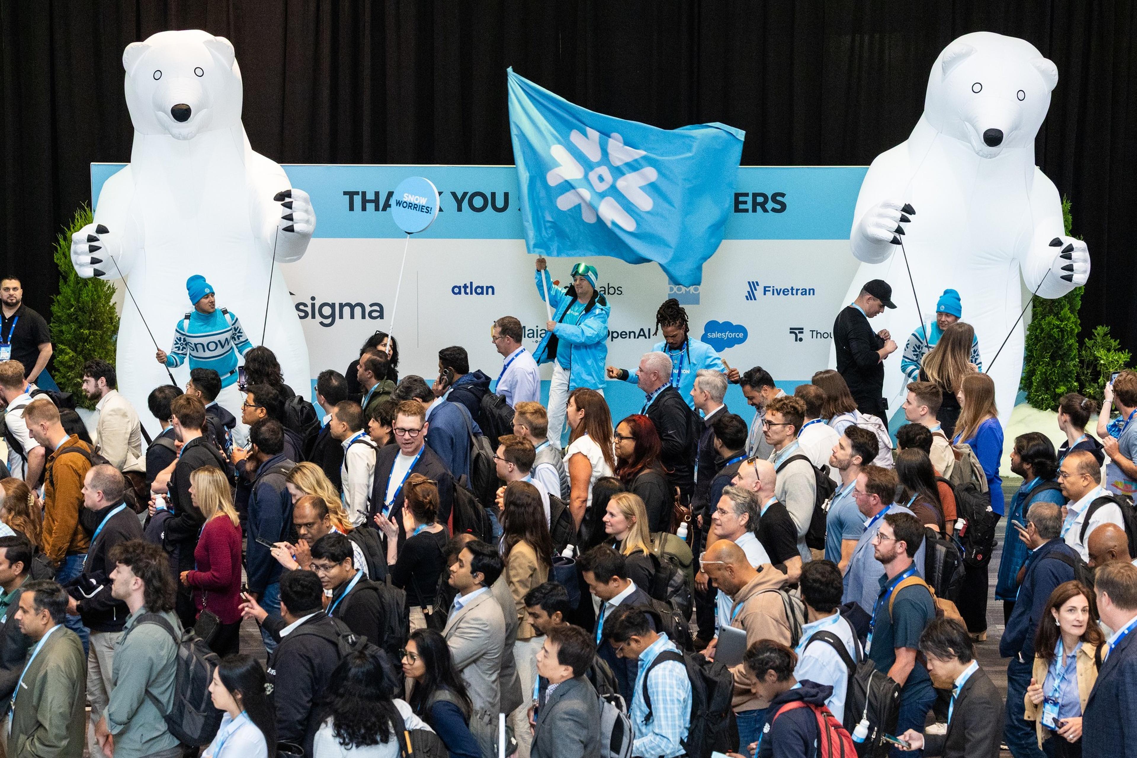 A crowded event with many people walking between two large inflatable polar bears, one person holding a blue flag with a snowflake design.
