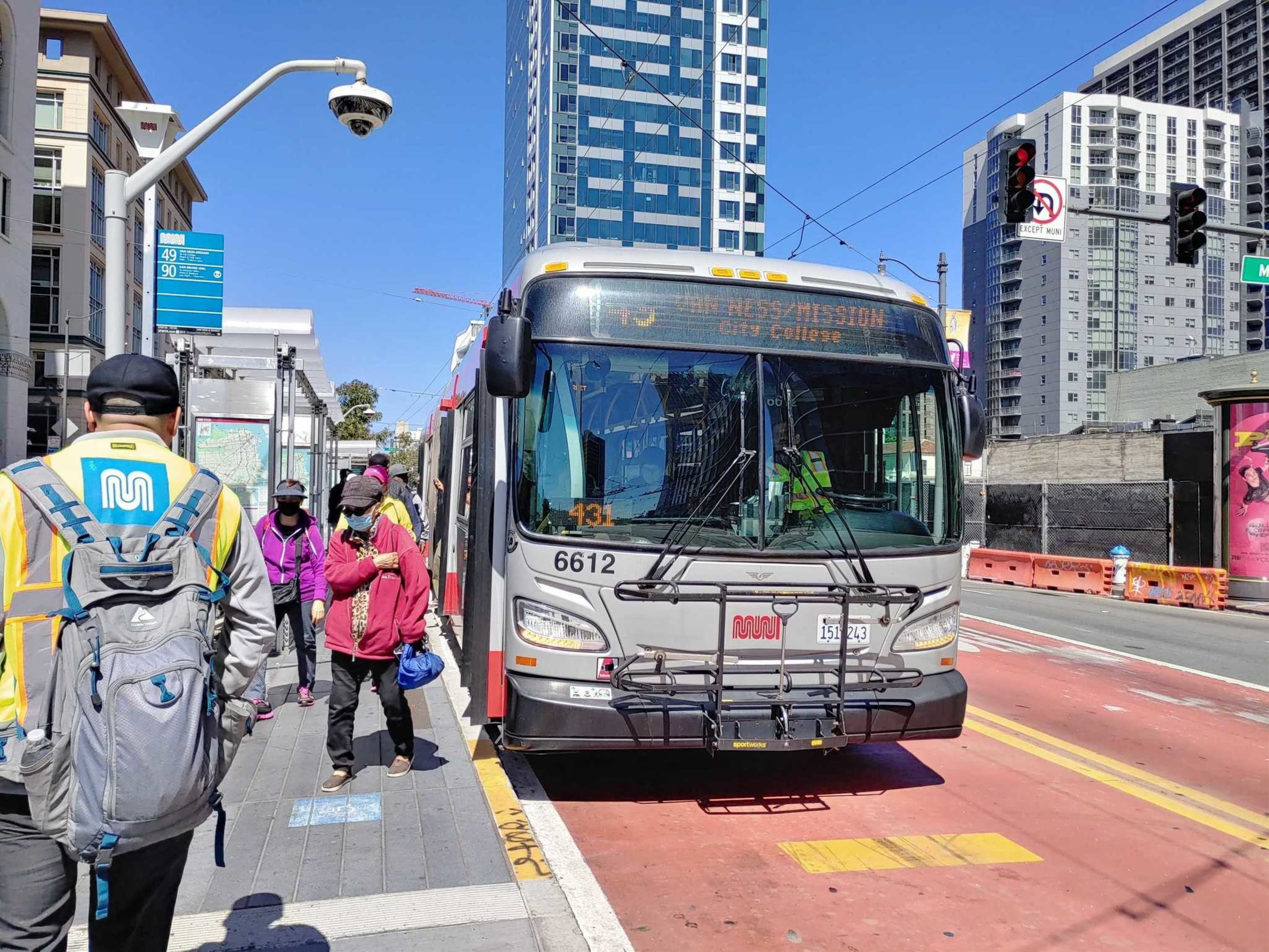 A Muni bus numbered 6612 waits at a stop on a sunny city street while people, some wearing masks, walk along the sidewalk next to tall buildings.