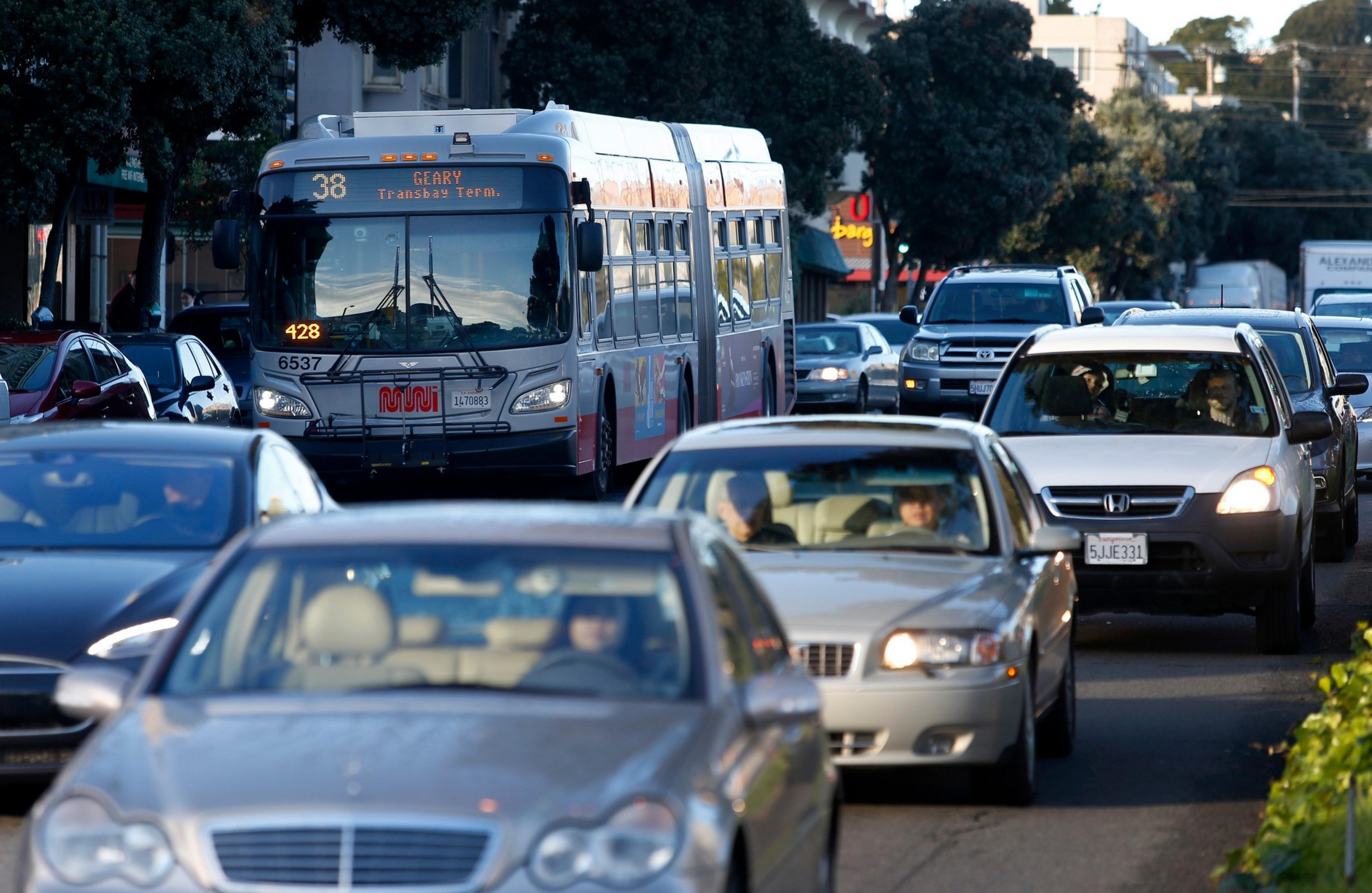 A city bus labeled “38 Geary Transbay Terminal” and several cars are in heavy traffic on a tree-lined urban street during daylight.