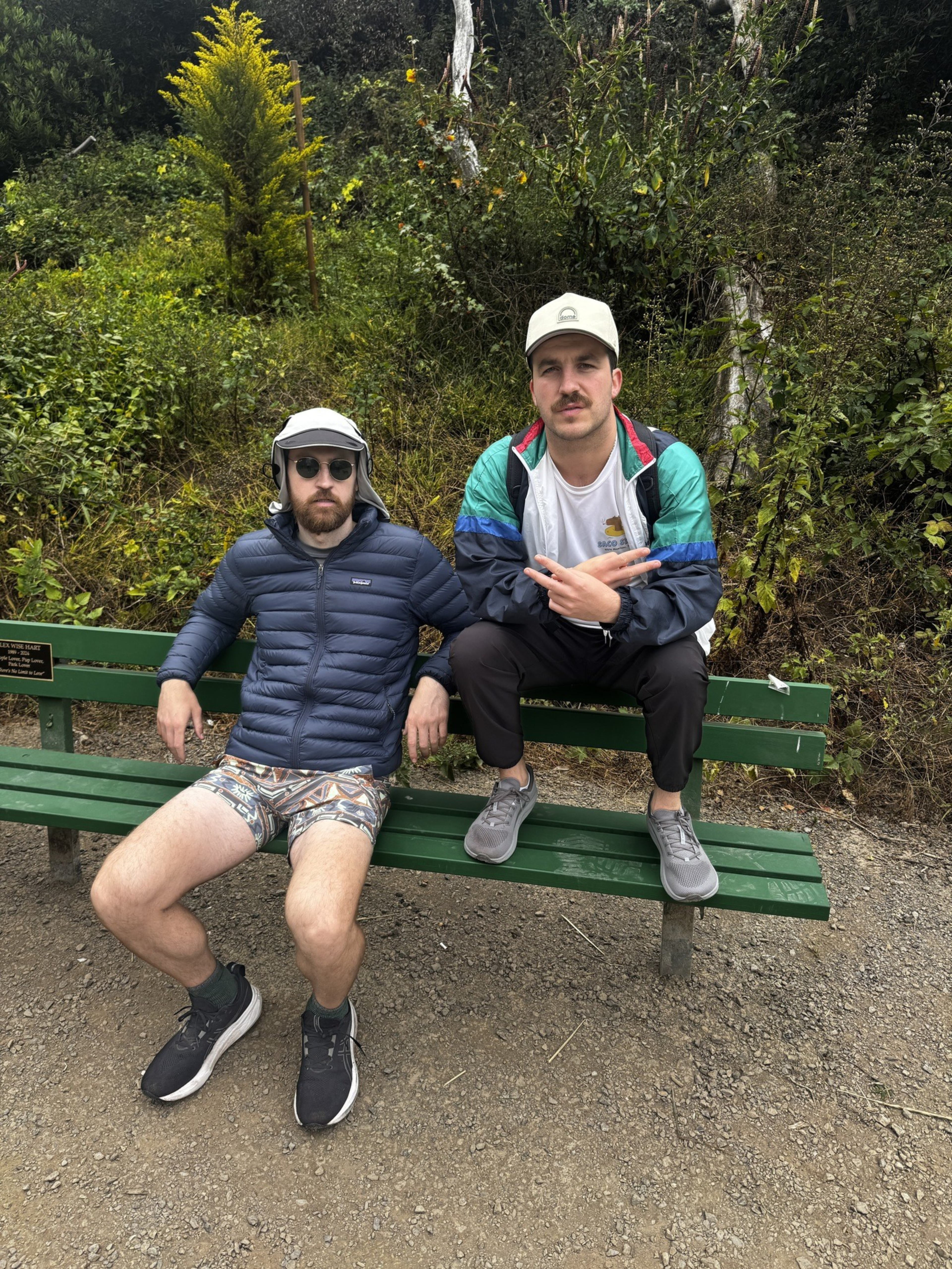 Two men in casual outdoor clothing sit on a green park bench surrounded by lush greenery. One wears sunglasses and the other flashes a peace sign.