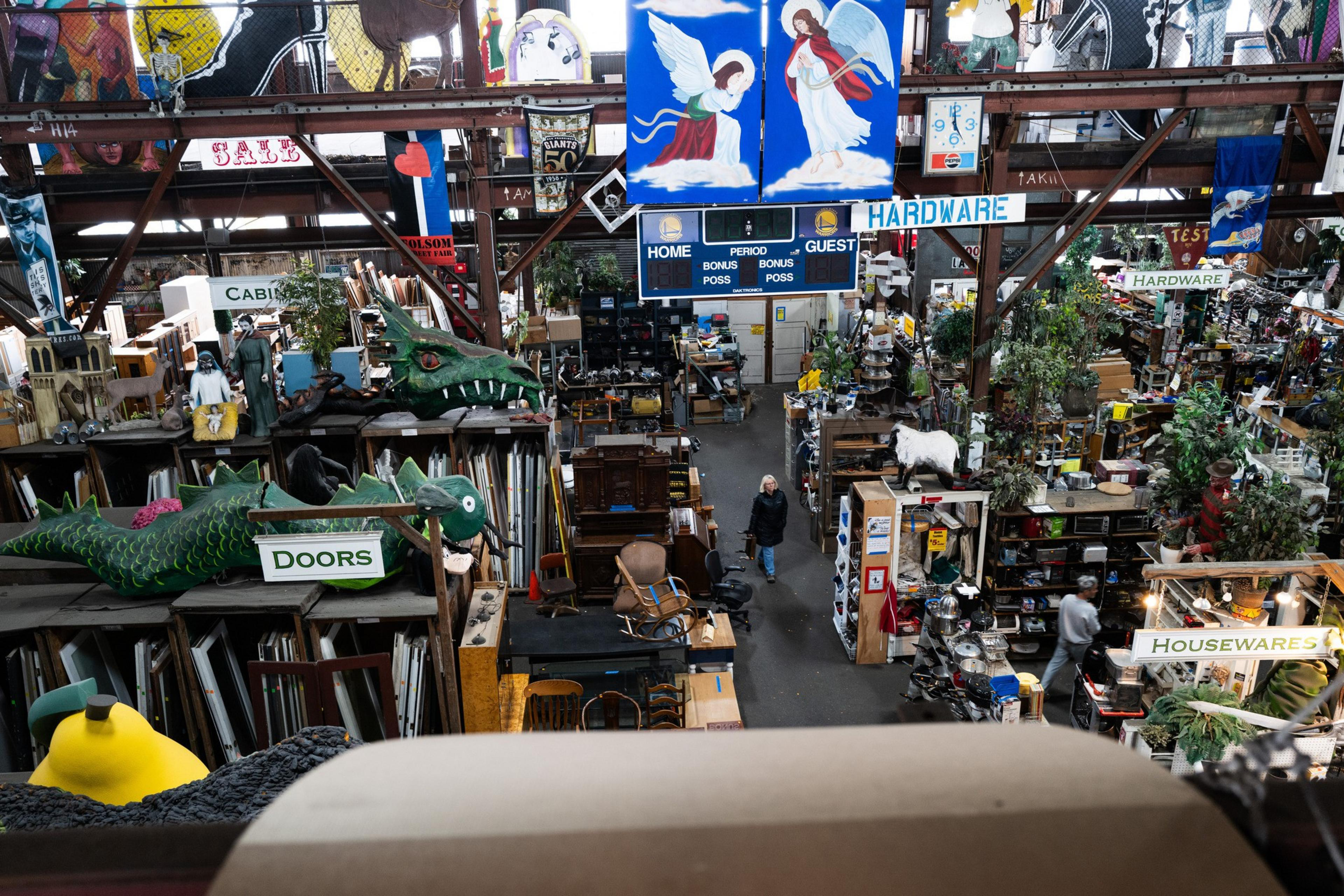 The image depicts a cluttered indoor warehouse filled with various items like doors, housewares, signs, and eclectic decorations. A scoreboard and banners hang above.