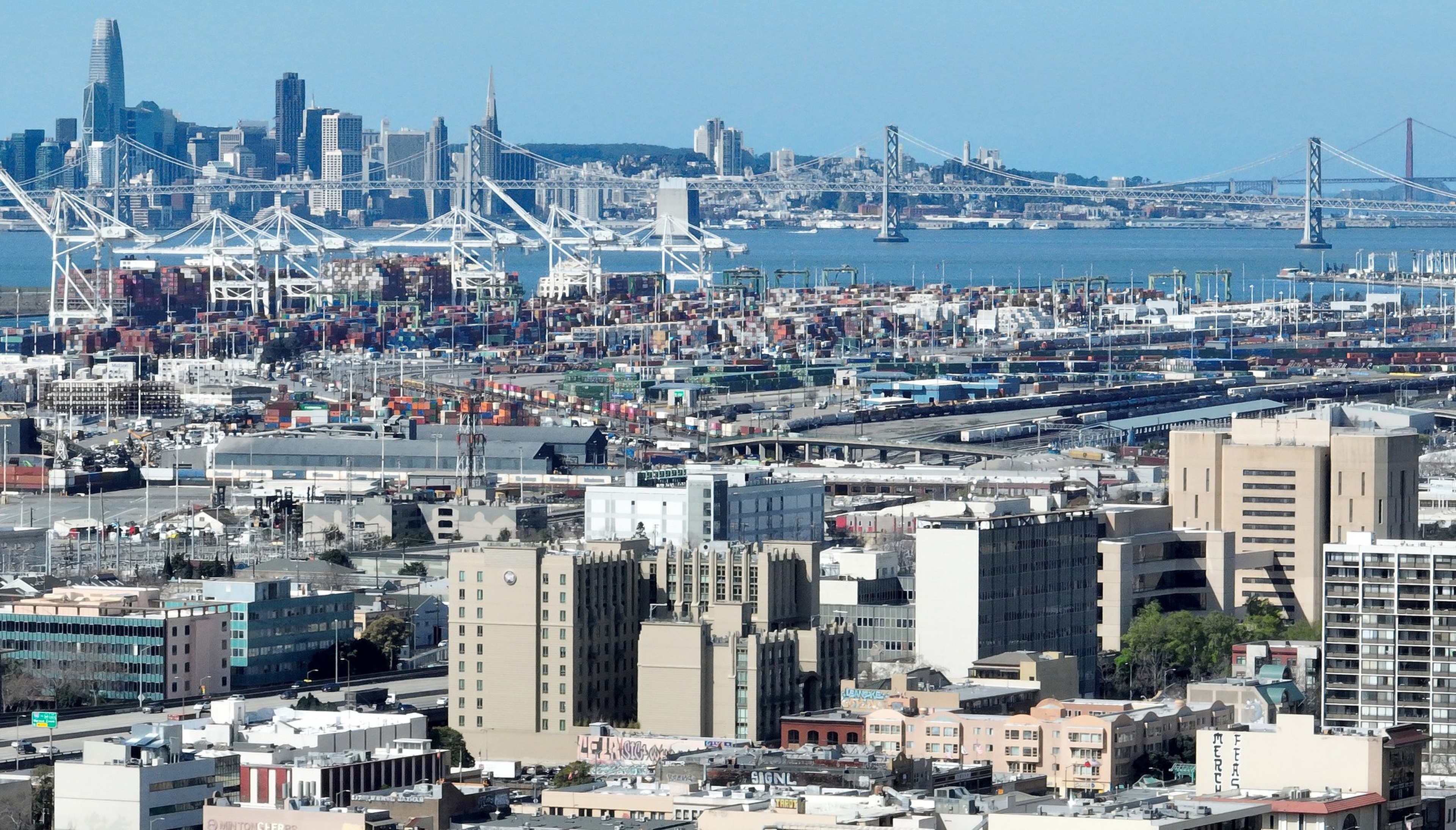A busy port with cranes and stacked shipping containers sits in front of a city skyline and a large suspension bridge over water under a clear sky.