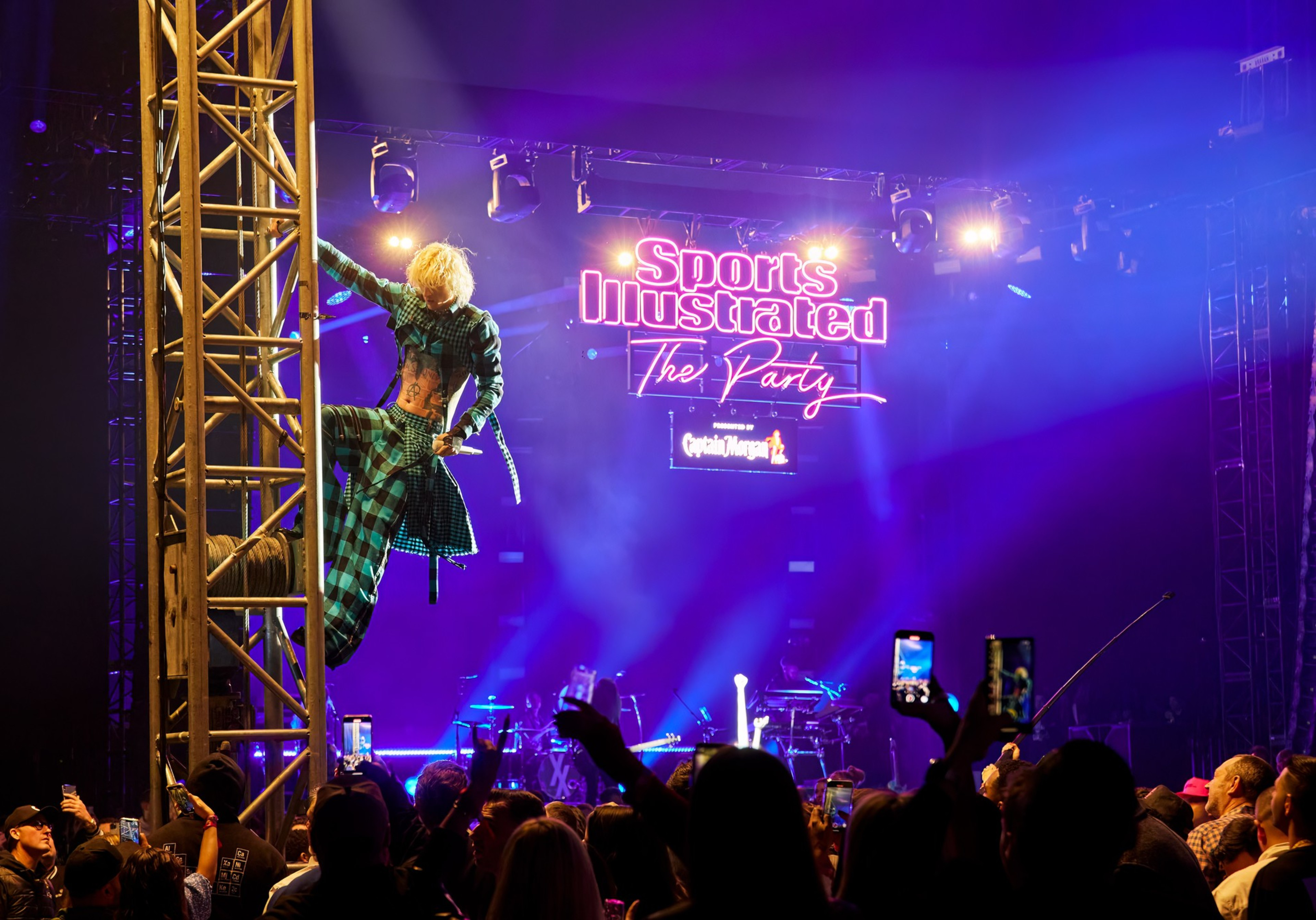 A performer in a plaid outfit climbs a metal structure on stage as a crowd watches and records under purple lights at a Sports Illustrated party.