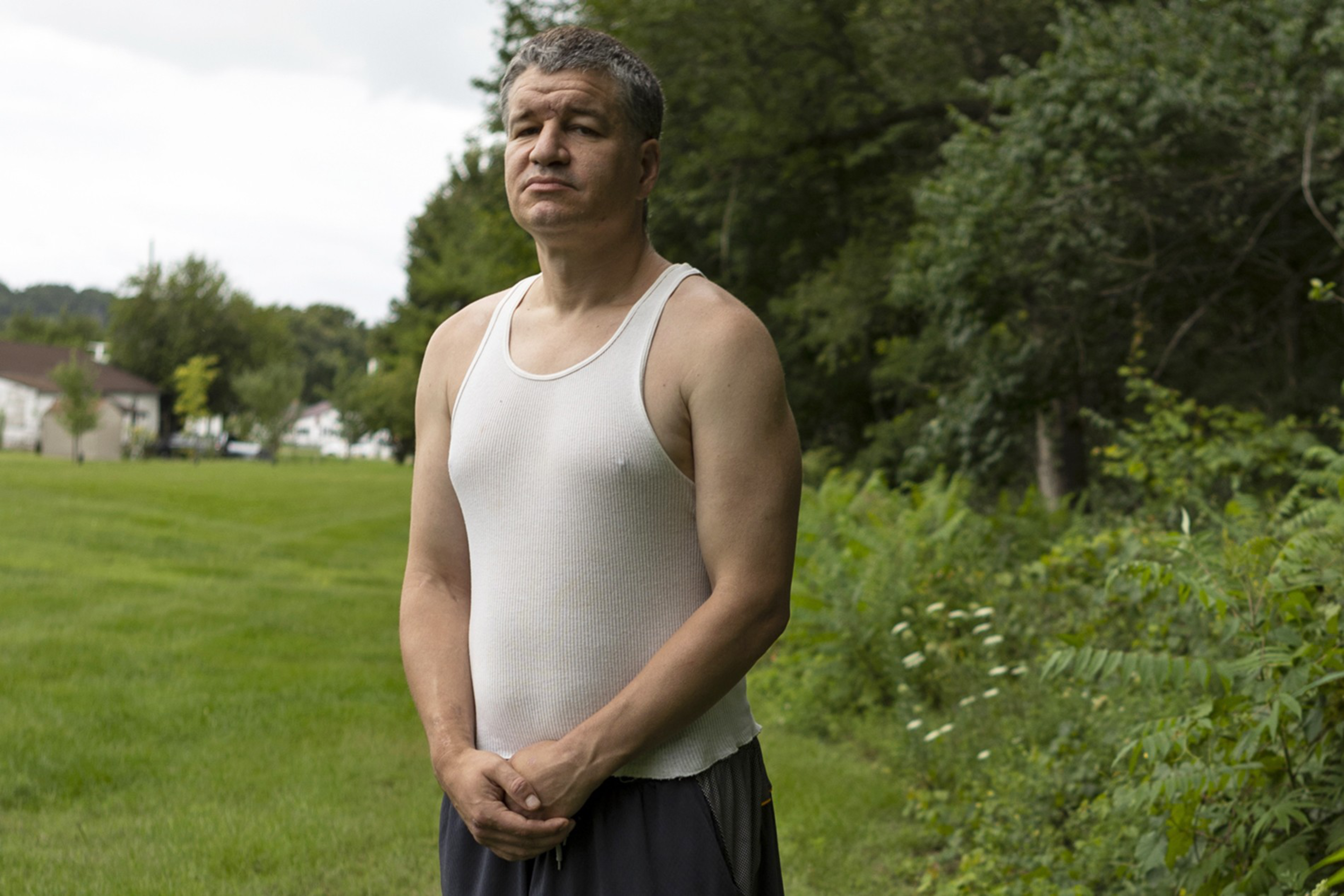 A man in a white sleeveless shirt and dark pants stands with folded hands on a grassy area near trees and houses in the background.