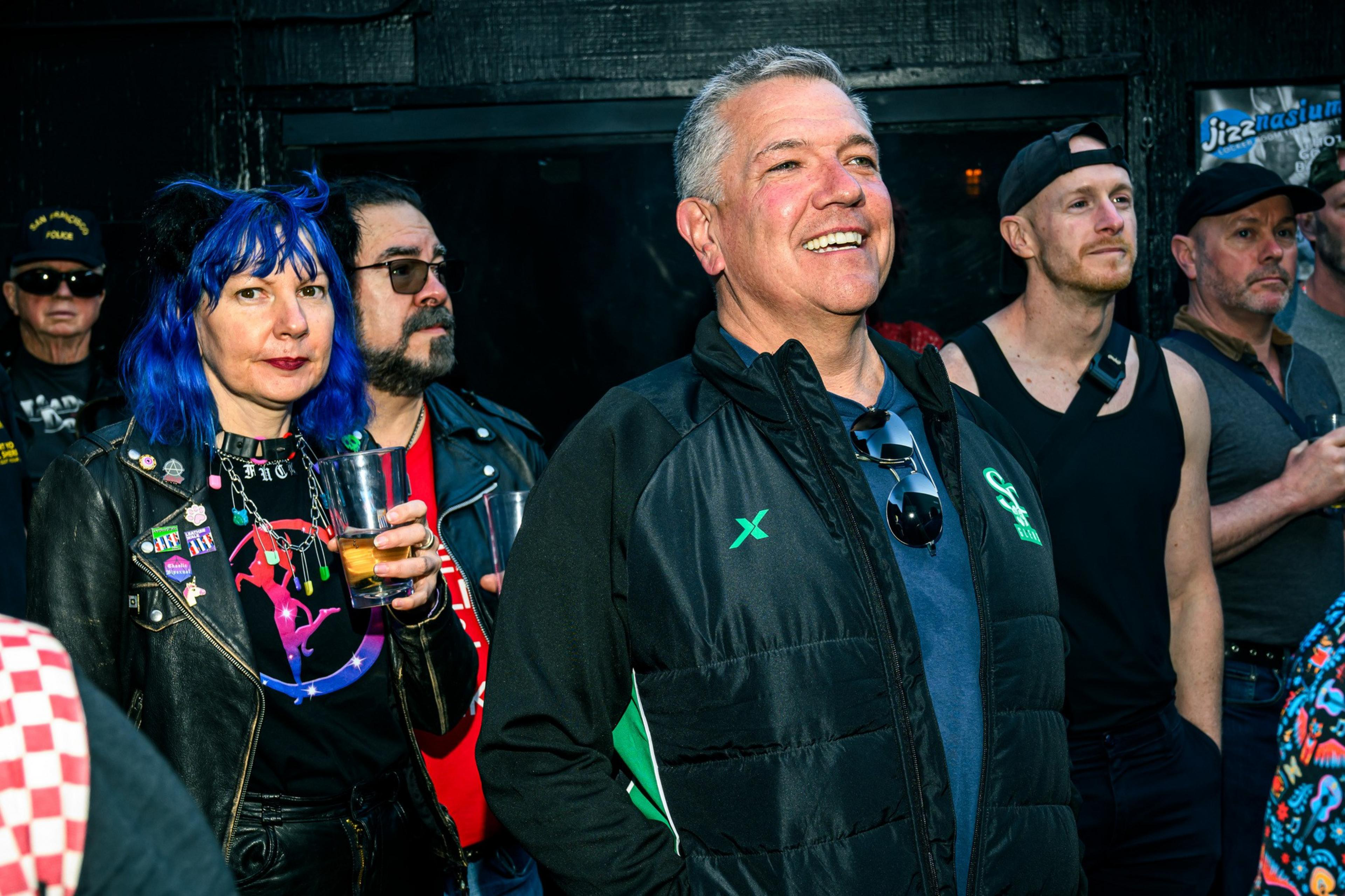 A group of people stands in a dimly lit area, smiling and holding drinks. One person has bright blue hair and is wearing a leather jacket with colorful pins.