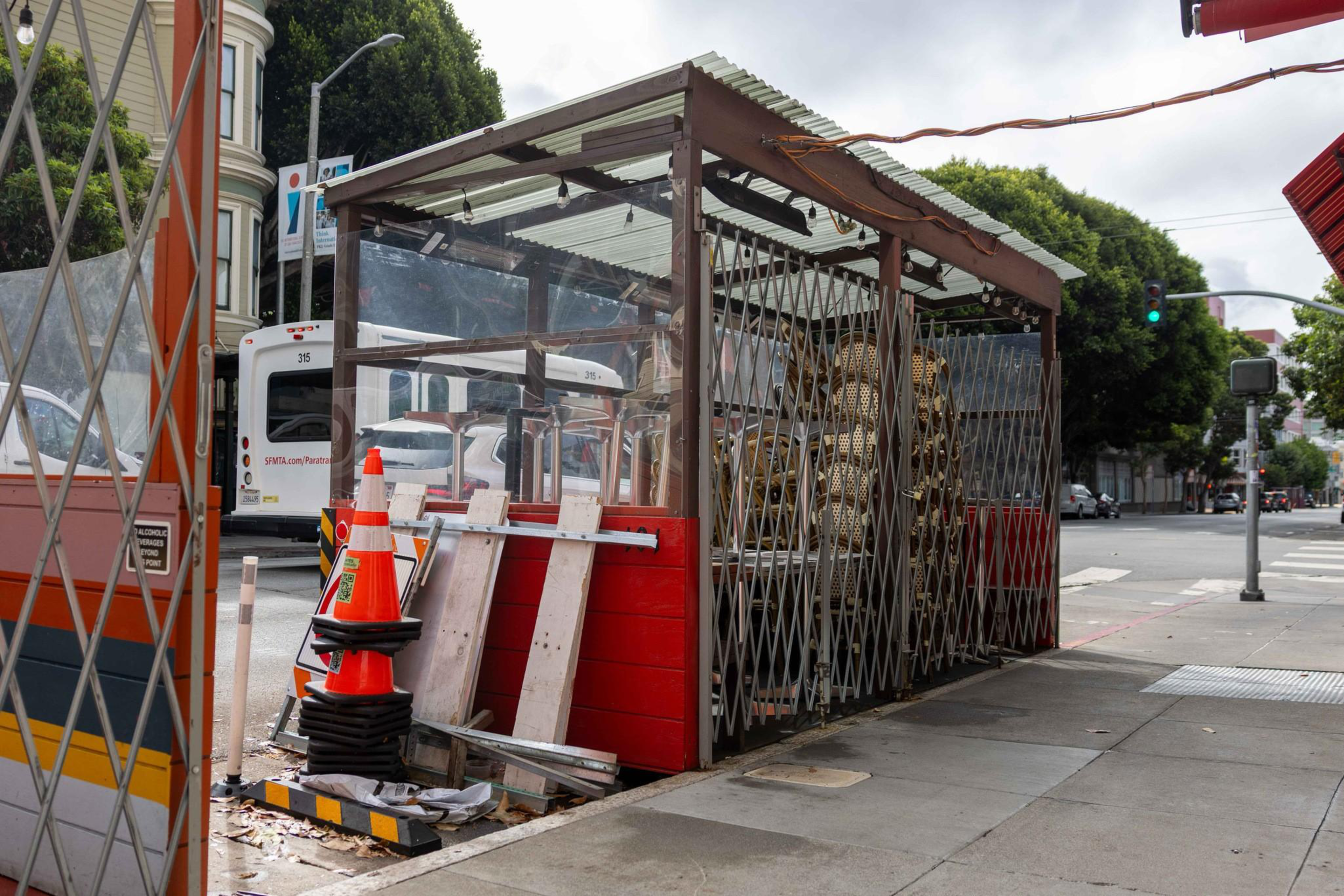 A small, red, temporary structure with metal gates houses stacked chairs beside a sidewalk, with a few traffic cones and a bus in the background.